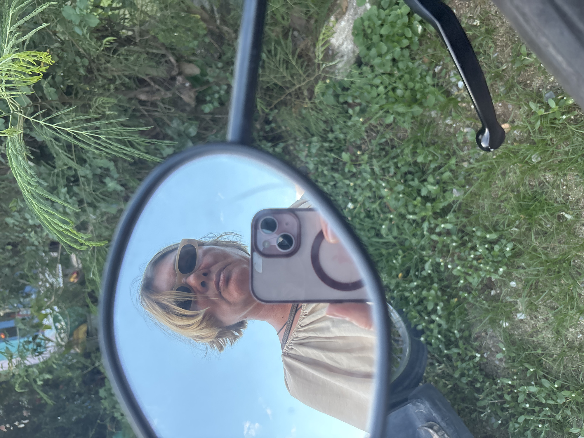 Captured through a motorcycle mirror, a woman with short blonde hair and sunglasses is taking a selfie with a phone. The mirror reflects the sky and nearby foliage. The scene is outdoors, in daylight. The background reveals green vegetation, suggesting a natural or park-like environment. A portion of the motorcycle, including the mirror's frame and handlebar, is visible. The location is Baan Bo Phut, Thailand.