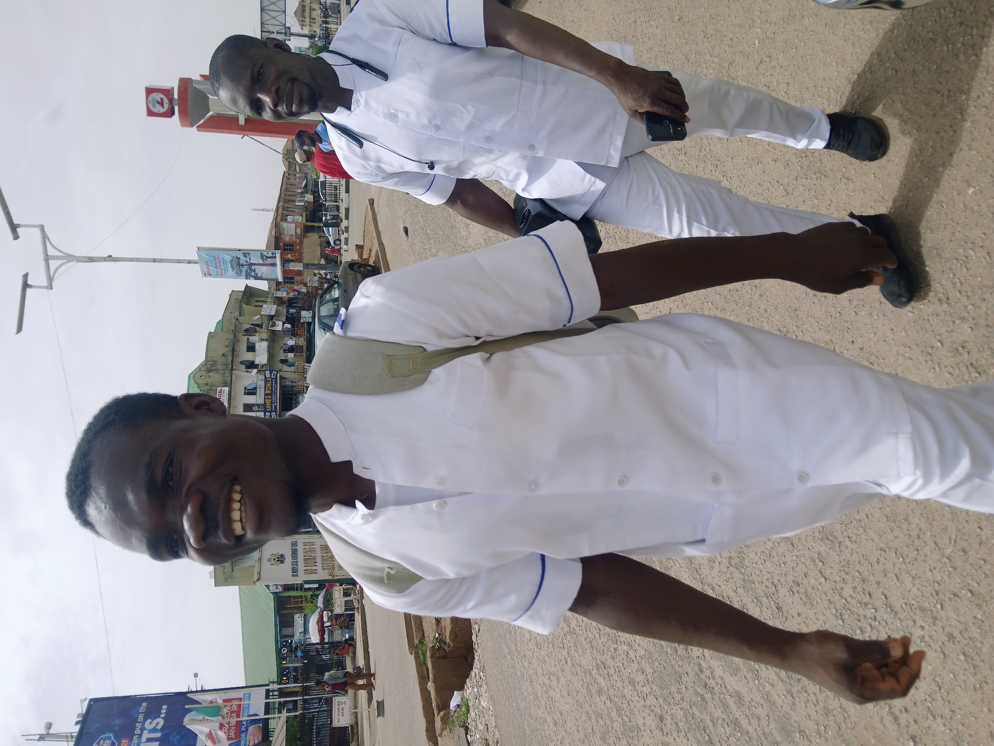 Two dark-skinned men in white uniforms with blue trim stand on a street in Jos, Nigeria. The man closest to the camera is smiling and looking directly at the camera, his right arm extended towards the viewer. A gray bag is slung over his shoulder. The other man is slightly behind him and also looking at the camera, holding a black smartphone in his right hand and wearing a stethoscope around his neck. Both men wear black shoes. The street is paved and appears to be in a commercial area, with buildings and signage visible in the background. A tall pole with light fixtures and a billboard for "ITS" are also visible. The billboard is signed by the Hon. Janearmigh, in Plateau State. Other signs are for Games World and are from the Jos North Local Government. The sky is overcast.