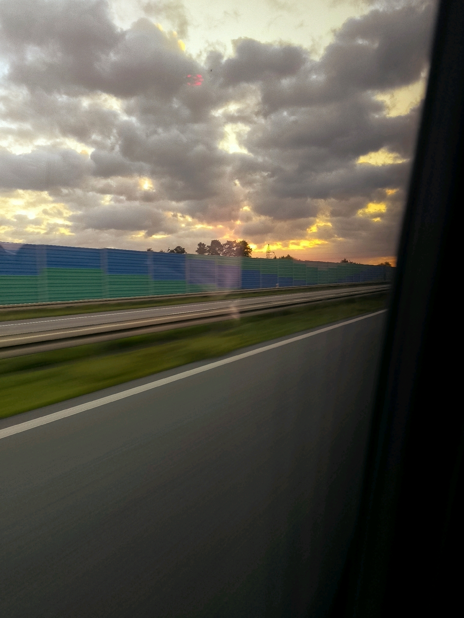 The image depicts a view from inside a vehicle traveling on a highway in Osiecza Druga, Poland. The road ahead is paved with asphalt and has white lane markings. On the left side of the highway, a sound barrier is visible, divided into sections of green and blue. Beyond the barrier, trees and a power line are silhouetted against a dramatic sky filled with dark, heavy clouds. The sun is setting, casting a warm, orange glow through the clouds, creating shafts of light. The motion of the vehicle is evident in the slight blur of the surrounding landscape. The right side of the image is dominated by a dark, out-of-focus area, likely the interior of the vehicle from which the photograph was taken.