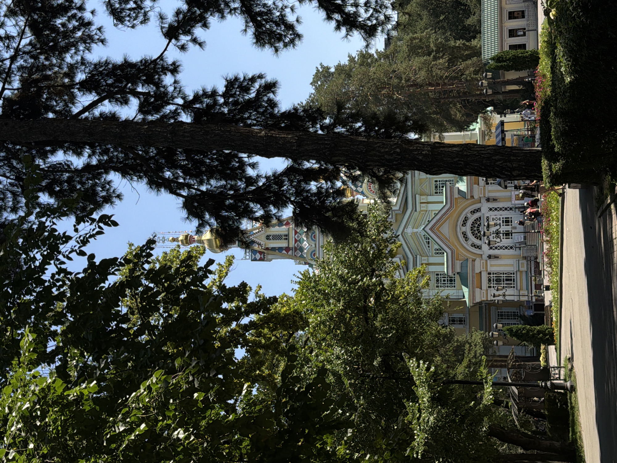The image shows the Zenkov Cathedral, a wooden Orthodox cathedral located in Panfilov Park in Almaty, Kazakhstan. The cathedral is brightly painted with a golden dome and intricate architectural details. It is surrounded by lush green trees, creating a park-like setting. Several people are visible in the image, walking around the cathedral and enjoying the park. The sky is a clear blue, suggesting a sunny day. The scene appears peaceful and scenic, with a mix of natural and architectural elements.