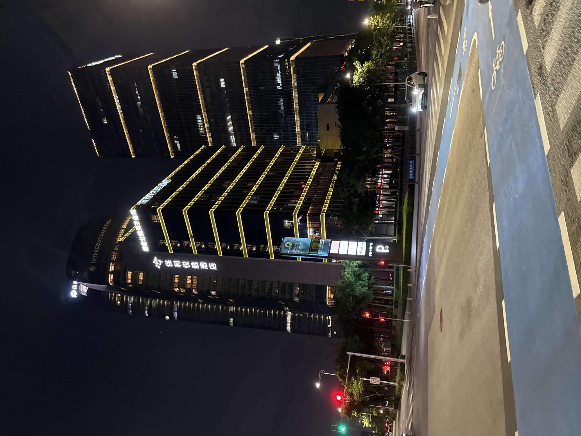 The image depicts an urban street scene at night in the Haizhu District of Guangzhou, China, featuring several illuminated high-rise buildings and road infrastructure.
In the background, three prominent skyscrapers dominate the skyline. The central building, partially curved, displays the illuminated "INTERCONTINENTAL" logo horizontally near its apex and a vertical sign in Chinese characters that reads "保利悦雅酒店" (Poly Yuya Hotel) down its facade. To its right, another tall, rectangular building is distinctly marked by multiple horizontal lines of bright yellow lights across its dark facade, creating a tiered effect. A shorter, multi-story building in the mid-ground also features similar horizontal yellow light strips, suggesting it is part of the same architectural complex. Further to the left, a third, taller skyscraper is partially visible, showing internal lights through its windows. Visible signage on the ground level, including a "P" for parking and text "保利中悦广场" (Poly Zhongyue Plaza), confirms the location as part of the Poly Zhongyue Plaza development, which houses the InterContinental Guangzhou Exhibition Center in Haizhu District.
The foreground is occupied by a wide, multi-lane asphalt road. White dashed and solid lines delineate traffic lanes and pedestrian crossings. A distinct bright blue bicycle lane, marked with white dashed lines and bicycle symbols, runs along the right side of the road, curving slightly to the right. On the left, a traffic light displays a green signal, while other traffic lights in the distance show red. Streetlights illuminate the road and surrounding areas.
Vegetation, primarily trees and shrubs, is planted along the median and sidewalks, casting shadows and highlights under the artificial light. One white, modern car with illuminated headlights is visible on the right side of the road, driving away from the viewer. A person on a bicycle or scooter is partially visible on the far right edge of the frame, near the blue bike lane. No other individuals are clearly discernible. The sky above is dark, indicating late evening or nighttime.
