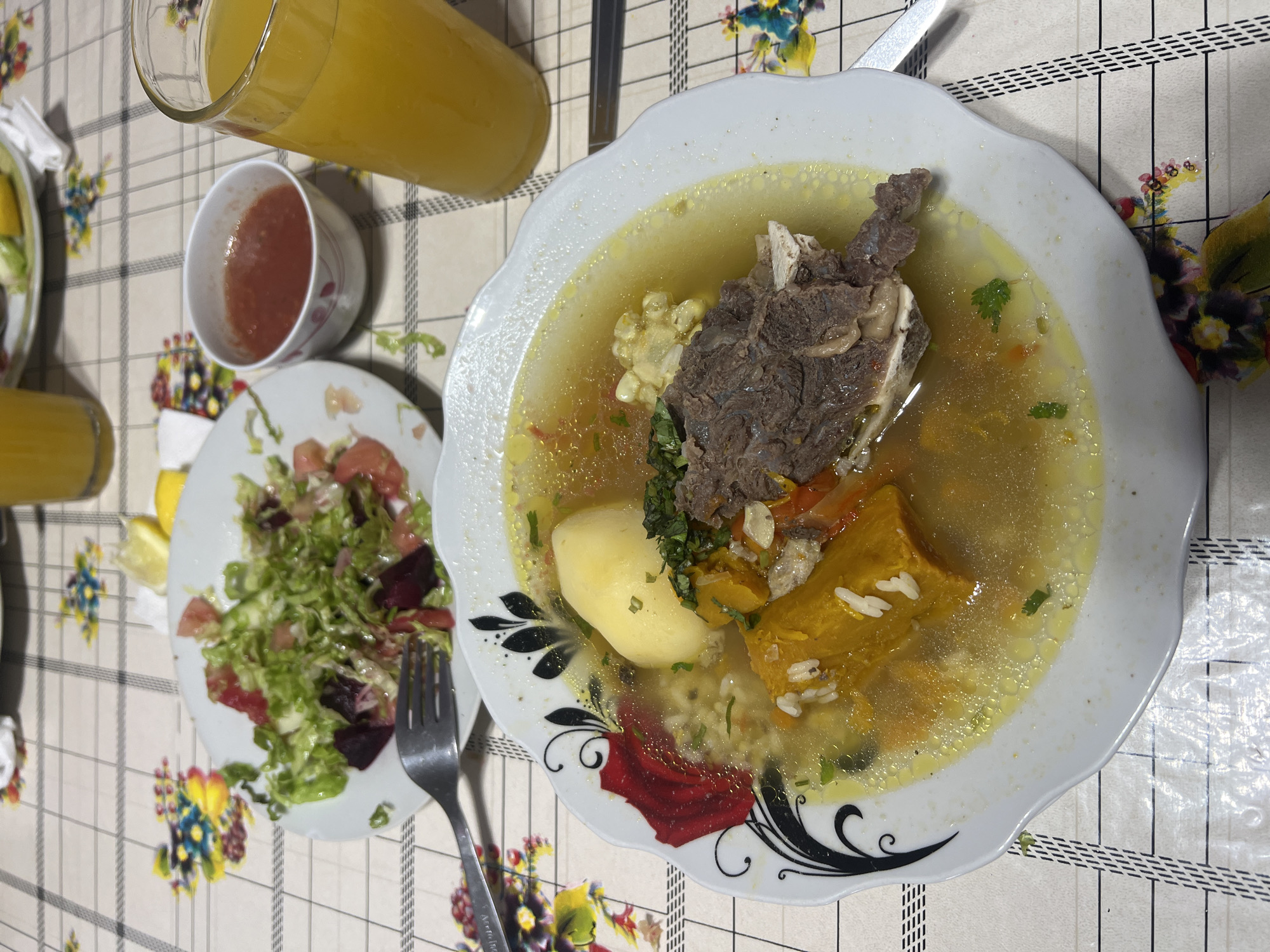 The image shows a close-up, overhead view of a meal served on a checkered tablecloth with floral accents. In the foreground, a white, scalloped-edge bowl contains a hearty soup. The soup appears to be a broth-based stew with visible chunks of meat, potato, corn on the cob, and orange squash or pumpkin, along with some rice and chopped green herbs. To the left of the soup bowl, a small white plate holds a salad of shredded lettuce, chopped tomatoes, and dark red pieces that could be beets. A fork rests on the edge of this plate. A small white bowl filled with a red sauce, possibly a salsa or relish, sits between the salad plate and a tall glass of orange juice. The glass of orange juice is positioned to the right of the frame, and another partially visible glass of orange juice is in the upper portion of the image. The lighting suggests an indoor setting, possibly a home or a casual restaurant, in Arica, Chile. There are no people visible in the image. The focus is on the food and the table setting.