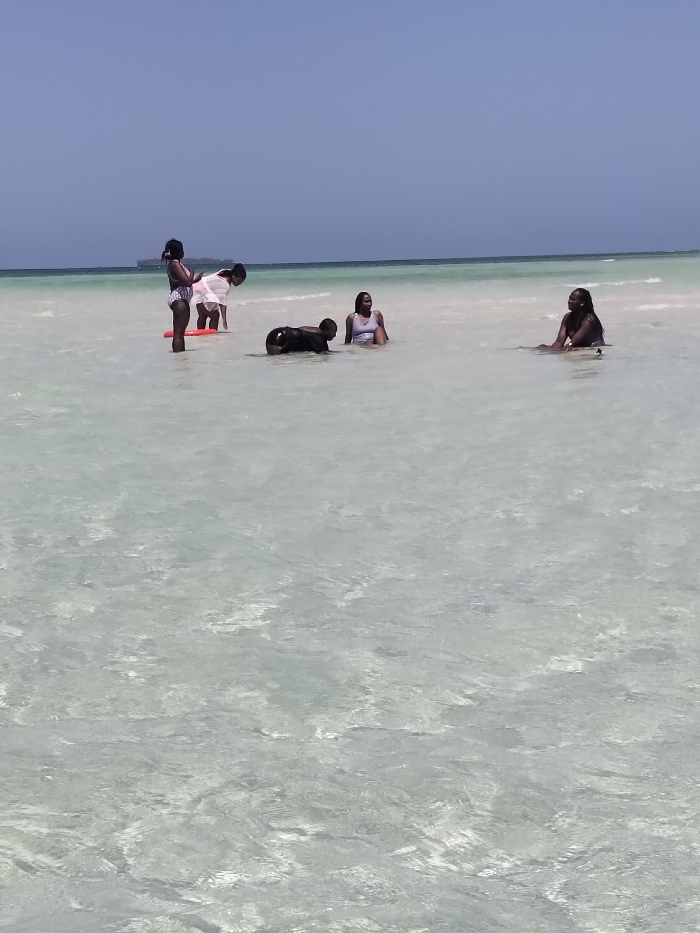 The image depicts five individuals in the shallow, clear, light blue waters off the coast of Wasini, Kenya, under a clear sky. From left to right: one individual stands, wearing a light-colored swimsuit, holding a red, rectangular flotation device. Behind them, a second individual in a white top and dark bottoms bends forward. In the center, a third individual, clad in dark clothing, is on all fours in the water. To their right, a fourth individual is seated in the water, wearing a light-colored swimsuit. Further to the right, a fifth individual is seated in the water, wearing a dark-colored swimsuit. The water extends to the horizon, where a faint, dark object, possibly a vessel or distant land, is visible. The scene is brightly lit by natural sunlight.