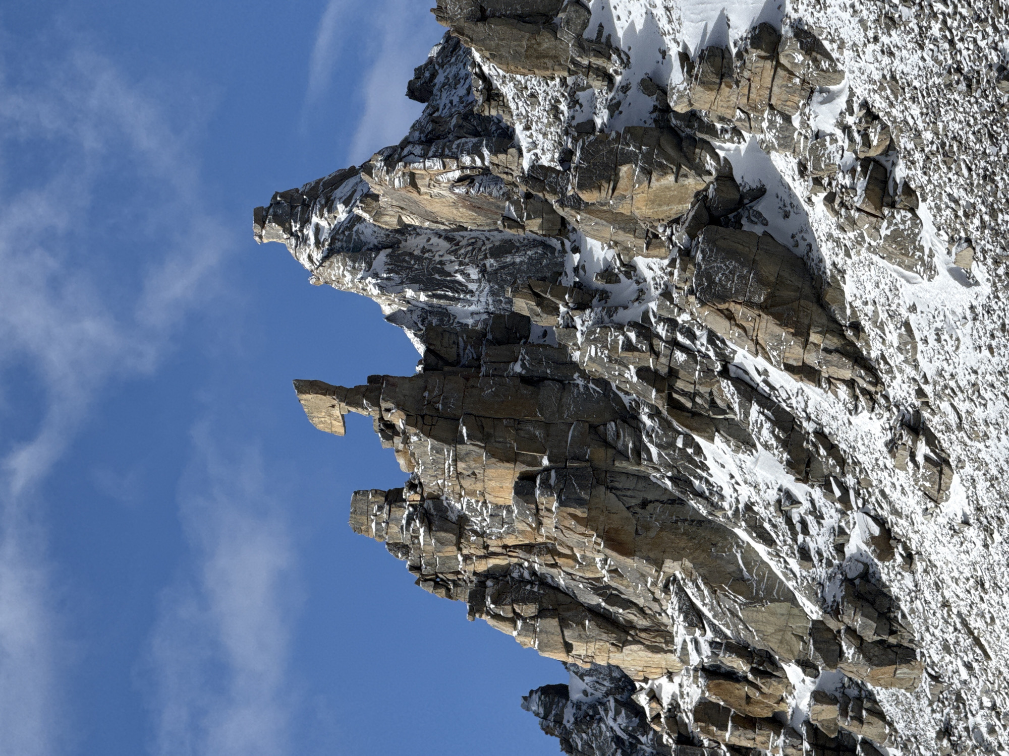 This image captures a dramatic, rugged mountain landscape, dominated by a steep, rocky peak and its upper slopes. The composition, when viewed with the sky at the top, features the mountain filling the lower, right, and central portions of the frame, while a clear blue sky with sparse, wispy clouds occupies the upper and left areas.
The mountain face is characterized by its deeply fractured, layered rock formations, displaying a palette of dark greys, browns, and hints of ochre. Patches of bright white snow cling to ledges, crevices, and gentler slopes, creating a stark contrast with the exposed rock. The texture of the rock is prominent, showcasing countless angular fragments and stratifications. A notable detail is a distinct, somewhat horizontal, flat-topped rock outcrop, resembling a 'nose' or a 'pinnacle', that projects outward from the main mountain mass into the sky.
The scene appears to be under bright sunlight, as indicated by the well-defined shadows that accentuate the rugged topography and depth of the rock face. The weather is clear and cold, allowing the snow to persist on the high-altitude terrain. There are no visible signs of people, animals, vegetation, or any discernible text within the frame. The image evokes a sense of grandeur, isolation, and the formidable power of natural landscapes.