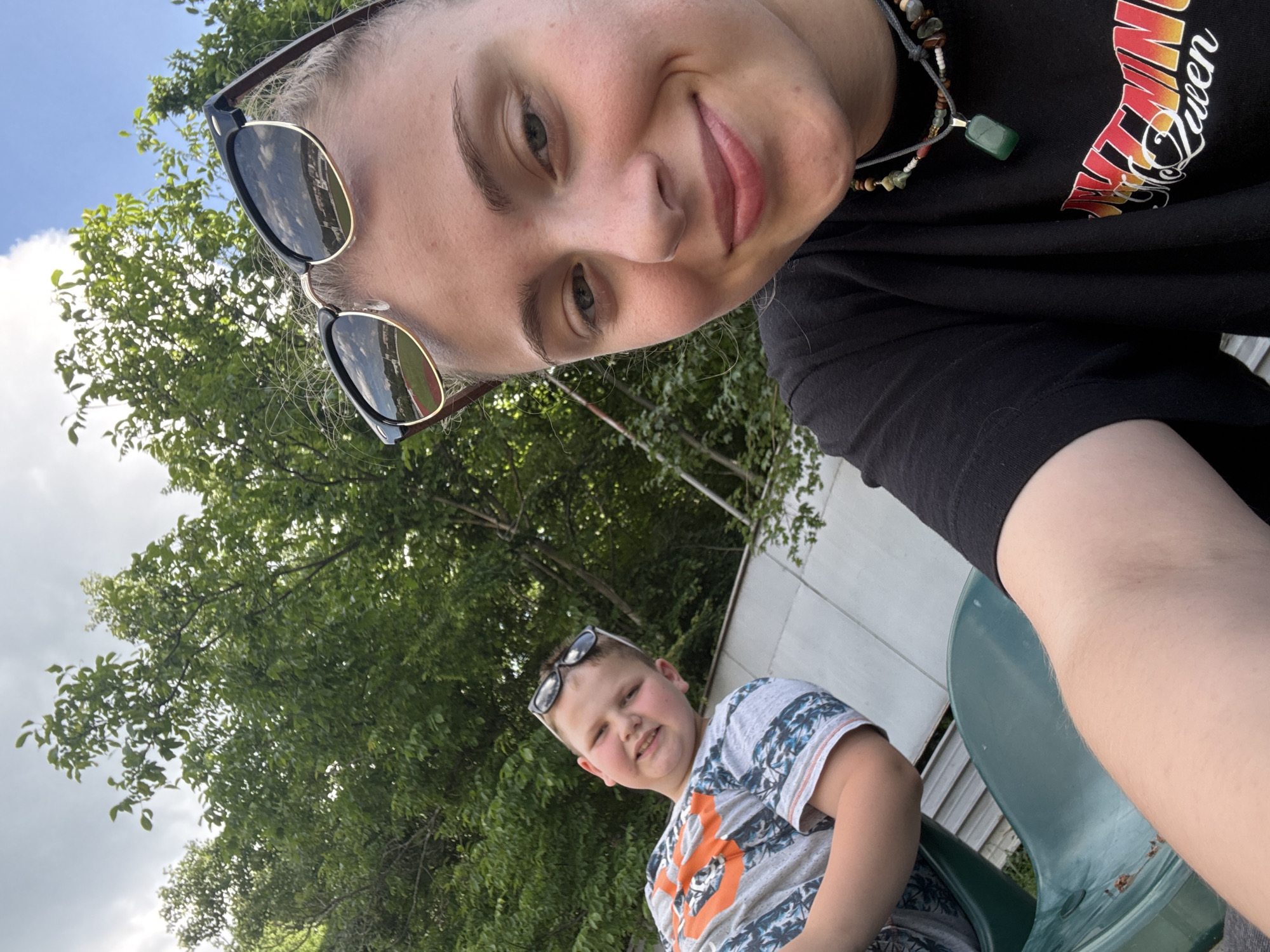 An outdoor, eye-level, medium shot shows two individuals sitting in green plastic stadium seats. The individual in the foreground, on the right, is a young woman with blonde hair pulled back, wearing sunglasses on top of her head, a black t-shirt, and a beaded necklace with a green pendant. She is looking towards the camera and smiling. The second individual, a young boy, is to the left and slightly behind her, wearing a grey and orange t-shirt and sunglasses on his head. He is also looking at the camera and smiling. Both are seated, and their arms are resting in their laps. Behind them, lush green trees are visible against a cloudy sky. The background appears to be an outdoor recreational area, possibly a park or sports field, given the stadium seating. The setting is consistent with an outdoor location in Gabrovo, Bulgaria, where such recreational facilities are common.