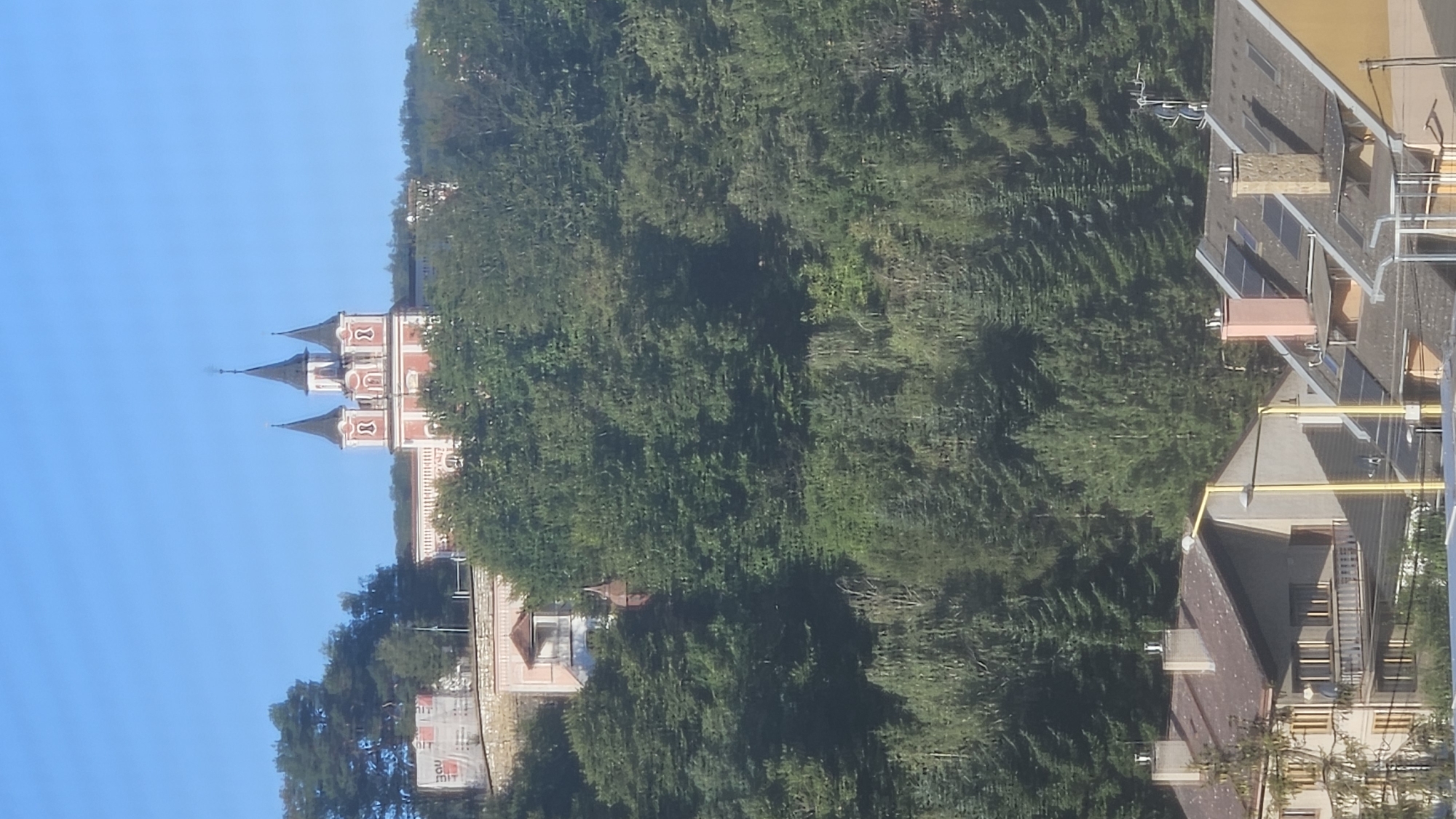 The image shows a view of a building with multiple spires, possibly a church or monastery, situated on a hillside covered in trees. The building features red and white architectural elements and appears to be constructed in a classical or baroque style. In the foreground, there are rooftops of residential buildings, suggesting an urban or suburban setting. The sky is clear and blue. The location is identified as the Solivar area of Prešov, Slovakia.