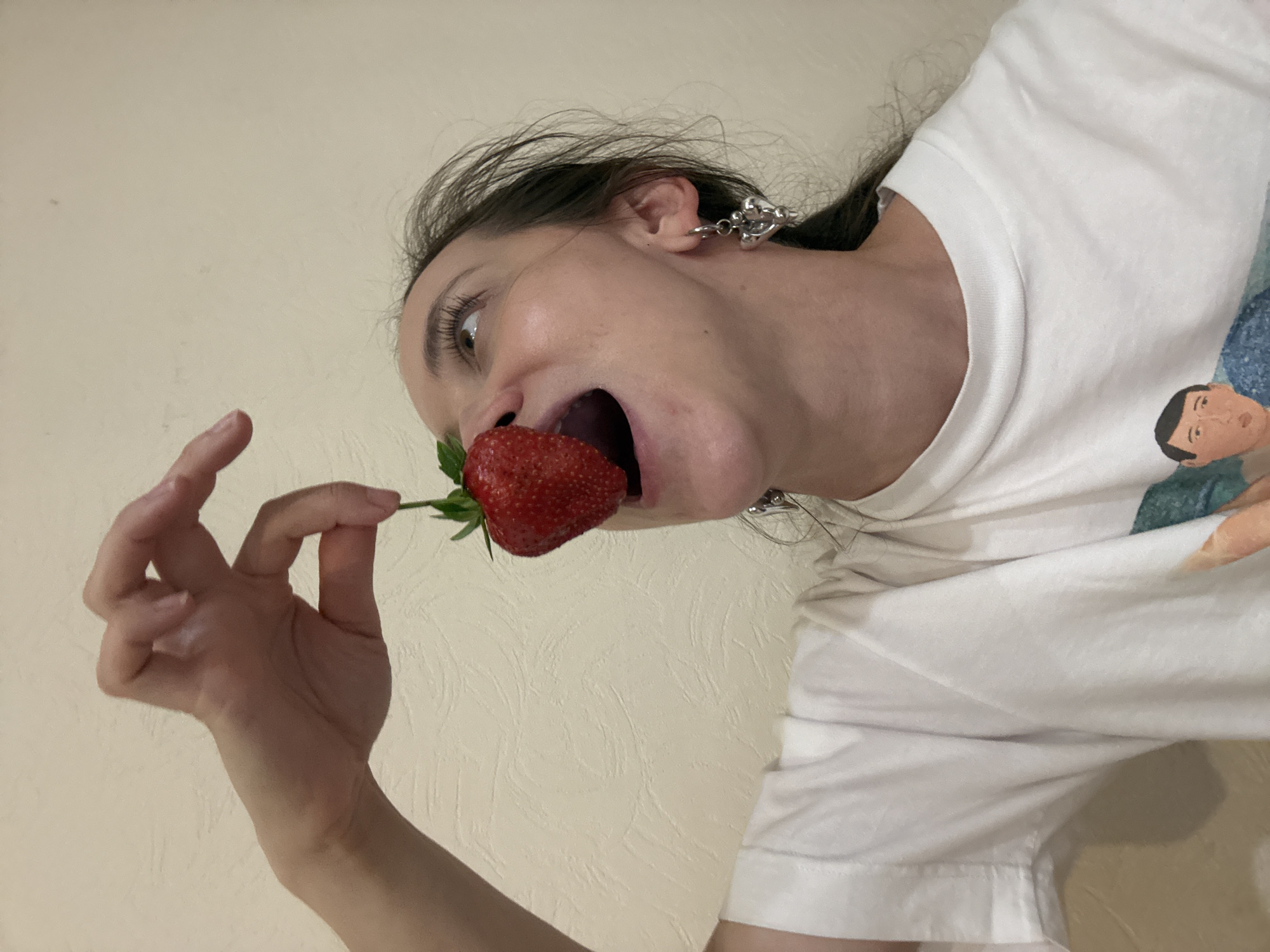 A person is holding a strawberry to their mouth. The person has light skin, dark hair, and is wearing silver earrings and a white t-shirt. The background is a plain, light-colored wall. There is no indication of the location being Kazan, Russia.