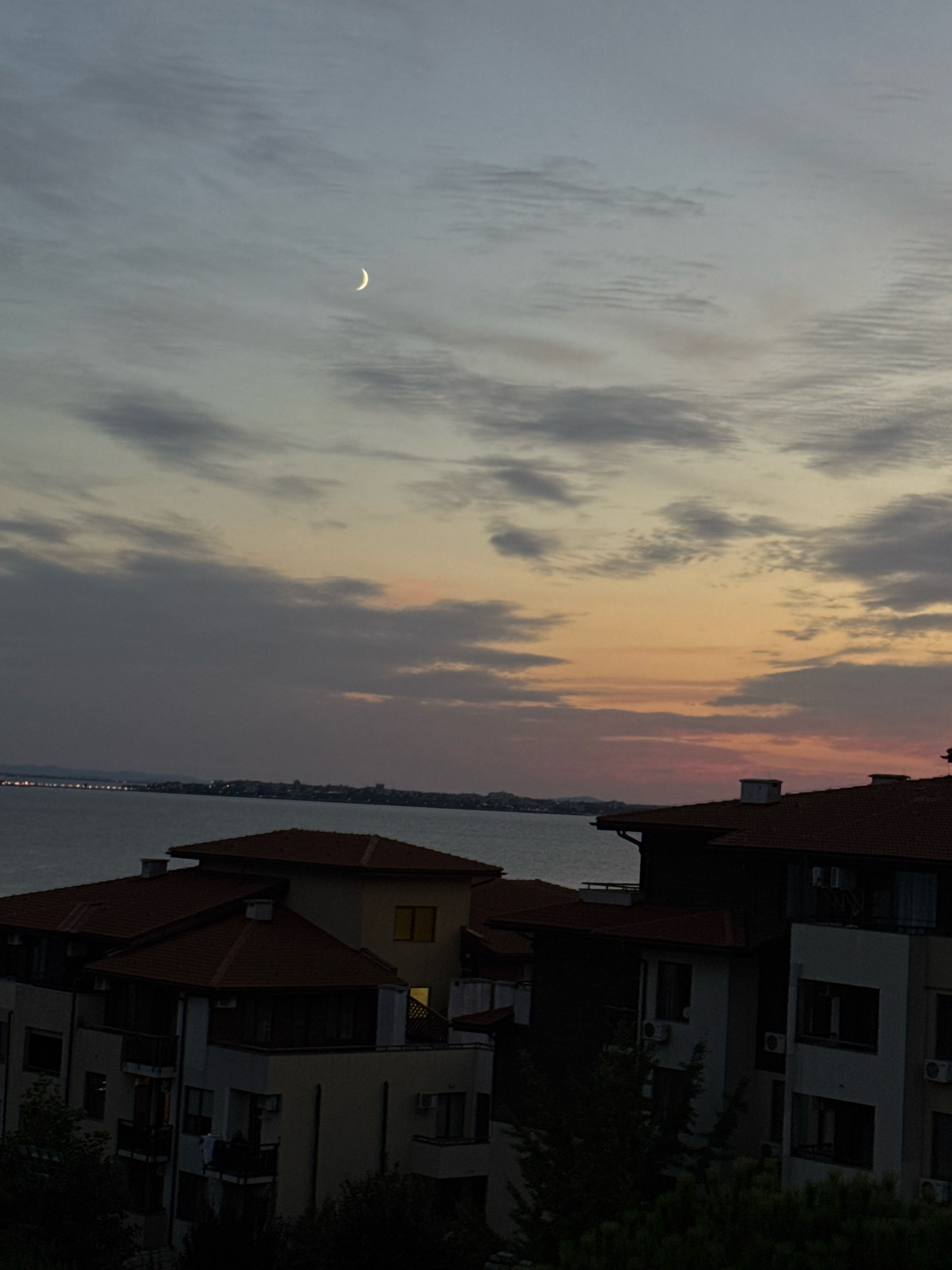The image depicts a twilight scene over the coastal city of Sveti Vlas, Bulgaria. A slender crescent moon is visible in the dusky sky, which transitions from pale blue at the top to hues of orange and pink near the horizon. Wispy clouds are scattered across the sky. Below the sky, a body of water, presumably the Black Sea, stretches across the frame. In the foreground, several buildings with terracotta tiled roofs are silhouetted against the fading light. The architecture suggests a residential or resort area, with balconies and multiple stories visible. Lights are beginning to appear in some of the building windows, indicating the transition from day to night. In the distance, the lights of the city are faintly visible along the coastline. The overall atmosphere is serene and tranquil.