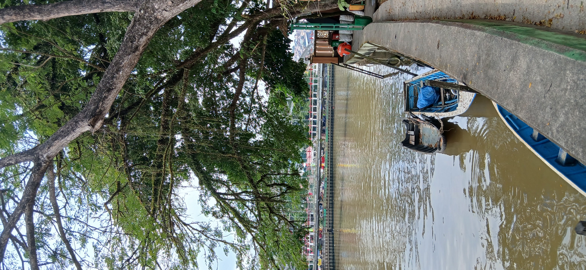 The image depicts the Mahakam River in Samarinda, Indonesia. Several small boats are moored along the concrete embankment. A large tree with dense foliage shades the foreground. In the background, a bridge spans the river, with buildings visible on the far bank. A person wearing an orange shirt is seated on the embankment, looking towards the river. The water is a murky brown, indicative of a river environment.
