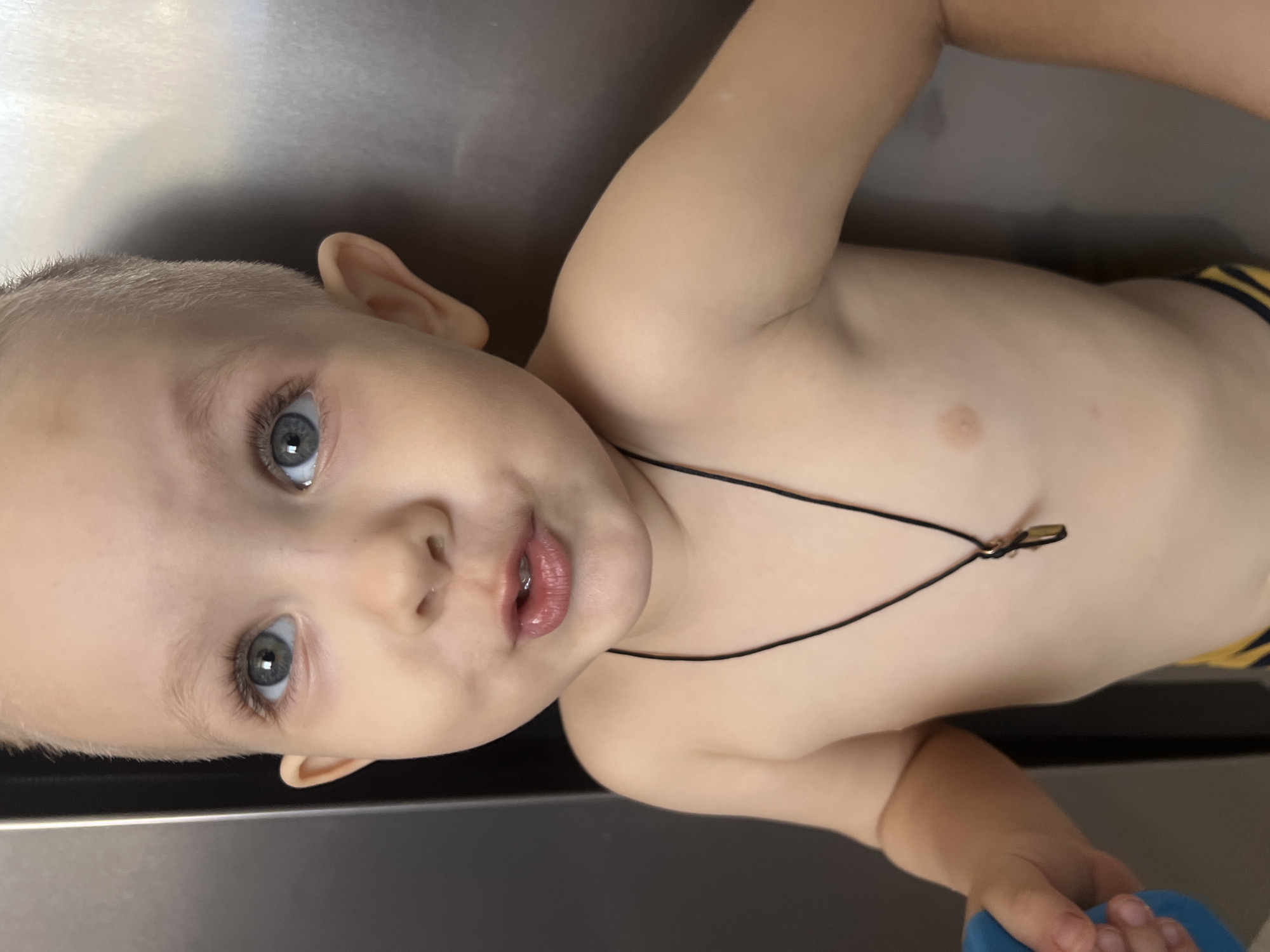 A close-up, slightly rotated image shows a young child lying on a smooth, grey surface. The child has light skin, blue eyes, and short, light-colored hair. The child's torso is visible and appears bare except for a thin, black cord with a small object attached, worn as a necklace. The child's right hand holds a blue object. The child is looking directly at the camera. The background is out of focus, but the surface the child is lying on appears to be flat and reflective. There is no indication of the city of Марковский, Russia in the image.