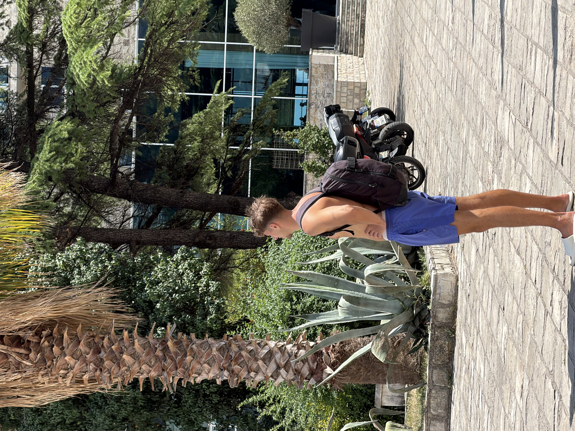 A young man wearing blue swim trunks and a black tank top stands on a paved walkway, his back to the camera. He has a black backpack slung over his shoulders. In the background, a modern building with a glass facade is visible, surrounded by lush green trees and a palm tree with dry fronds. A motorcycle is parked nearby. The scene appears to be outdoors on a sunny day, with the bright sunlight casting shadows. The man is looking down, and his posture suggests he might be resting or observing something on the ground. The overall setting is a resort or a hotel with a landscaped garden, likely in a warm climate. The location is specified as Budva, Montenegro.
