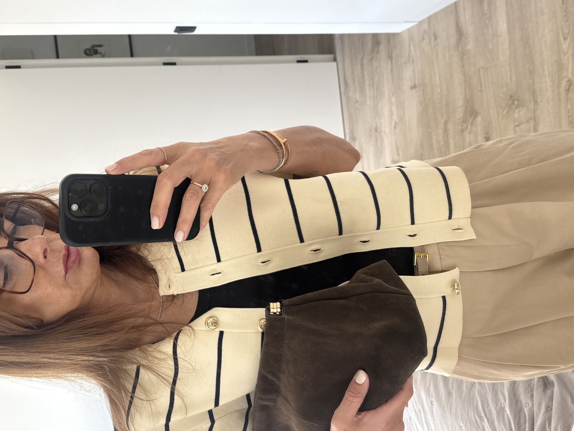 A person is taking a selfie in a room with light-colored wooden floors and white walls. The person is wearing glasses, a cream-colored cardigan with black stripes and gold buttons, and a black top underneath. They are holding a black smartphone with multiple lenses, showcasing jewelry on their fingers and wrist, including rings and bracelets. Part of a brown suede purse is visible in their other hand. Vila Nova de Gaia, Portugal is not discernible from the image.