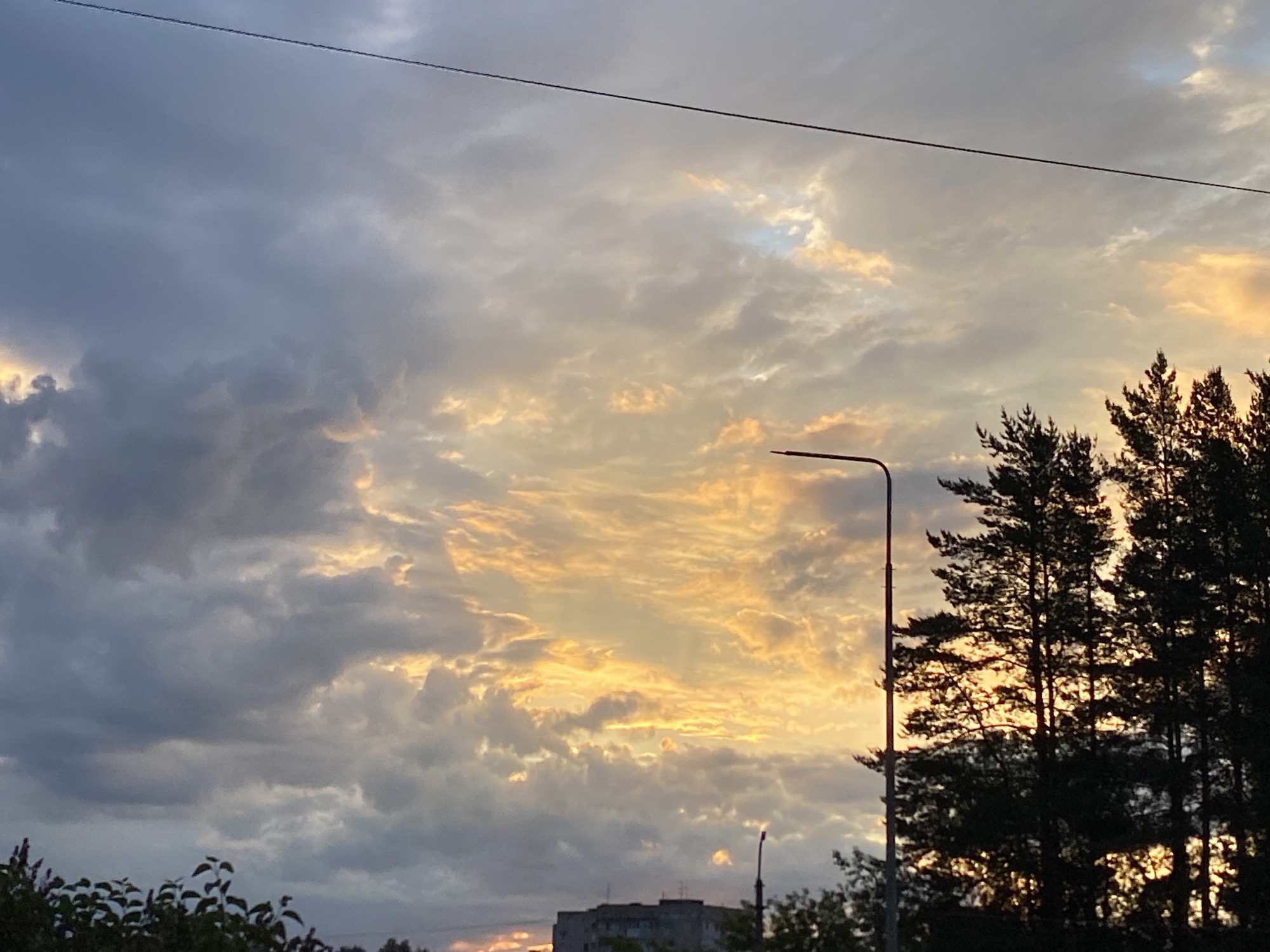 The image displays a dramatic sky at either sunrise or sunset over what appears to be an urban or suburban area in Chaikovsky, Russia. The sky is a prominent feature, showcasing a striking contrast between dense, dark grey clouds on the left and vibrant orange and yellow clouds in the center and right, indicating the low angle of the sun. Patches of lighter, possibly blue sky are visible through breaks in the cloud cover.
In the mid-ground, a tall street light pole stands on the right side, silhouetted against the bright clouds. To its right, a group of coniferous trees, likely pines, appears as dark silhouettes, their textured branches contrasting with the illuminated sky. A thin, dark power line or cable stretches horizontally across the top quarter of the frame.
Towards the bottom center, a multi-story building, possibly a residential block, is partially visible in silhouette, its structure hinted at through the foliage in the immediate foreground. The overall impression is one of a serene moment during dawn or dusk, with natural elements like trees and clouds coexisting with urban infrastructure. No people or specific activities are depicted. No text is visible in the image.