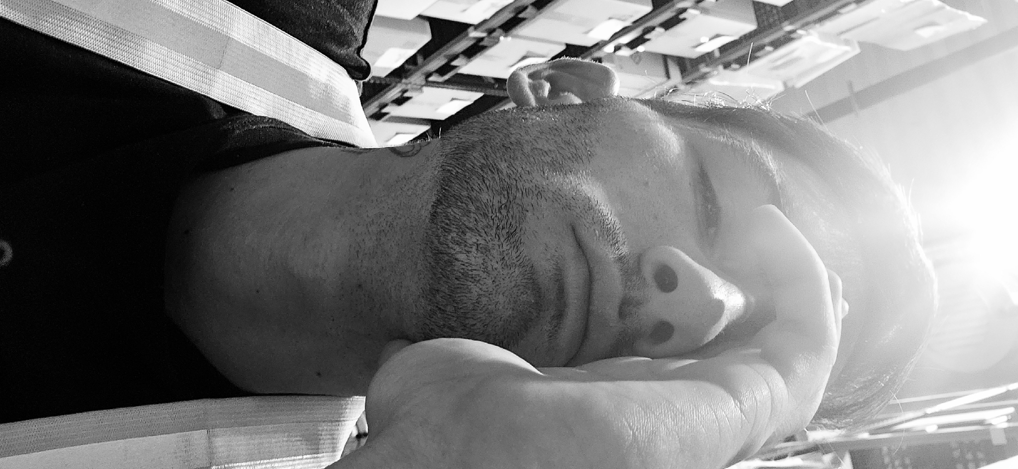 A close-up, black and white photograph depicts a man resting his face on his hand. The man has a trimmed beard and his eyes are closed. His head is angled, placing his ear towards the top of the frame.
The man is wearing a dark shirt with a light-colored, textured stripe across the shoulder. The background is out of focus, but appears to be a ceiling structure with repeating rectangular shapes and light fixtures. The intense source of light on the right side of the image casts flares.
The exact location in Plovdiv, Bulgaria is impossible to determine from the given image. There are no discernible landmarks or features in the photograph that are uniquely identifiable to that specific city.