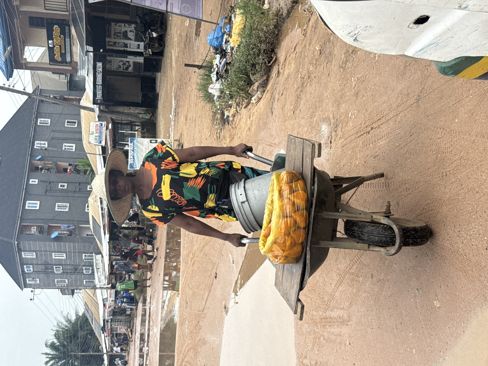 In Ihiagwa, Nigeria, a woman wearing a wide-brimmed straw hat and a colorful, abstract-patterned shirt pushes a wheelbarrow laden with a large bag of oranges and a grey bucket. The wheelbarrow has a wooden plank placed across its top, supporting the orange bag and bucket. The woman's left hand grips the wheelbarrow's handle. The ground is a muddy, reddish-brown dirt road with several large puddles reflecting the overcast sky. In the background, buildings line the street, some with businesses like "VERAP AUL COMMUNICATION" and "PEERLESS BARBING SALON" visible. Trash and vegetation are piled along the side of the road. Several people are visible in the distance, walking or standing near the shops. A white vehicle is partially visible in the foreground on the right.