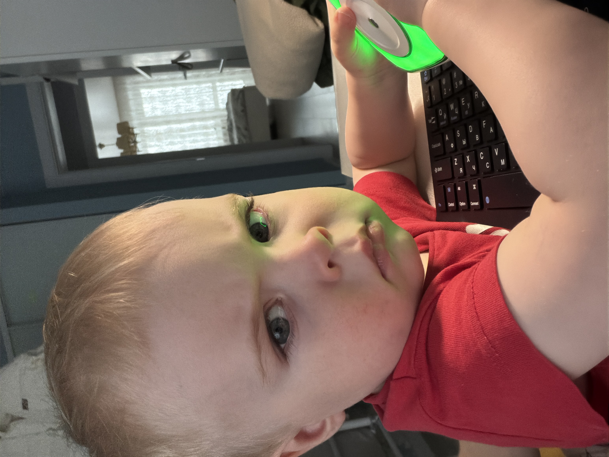 A baby lies on his back, holding a glowing green object with a white center above his face. The baby is wearing a red shirt. A black keyboard is visible near the baby's hands. The background includes a cabinet, a hanging bag or pillow, and a window with blinds. The location is undeterminable from the image.