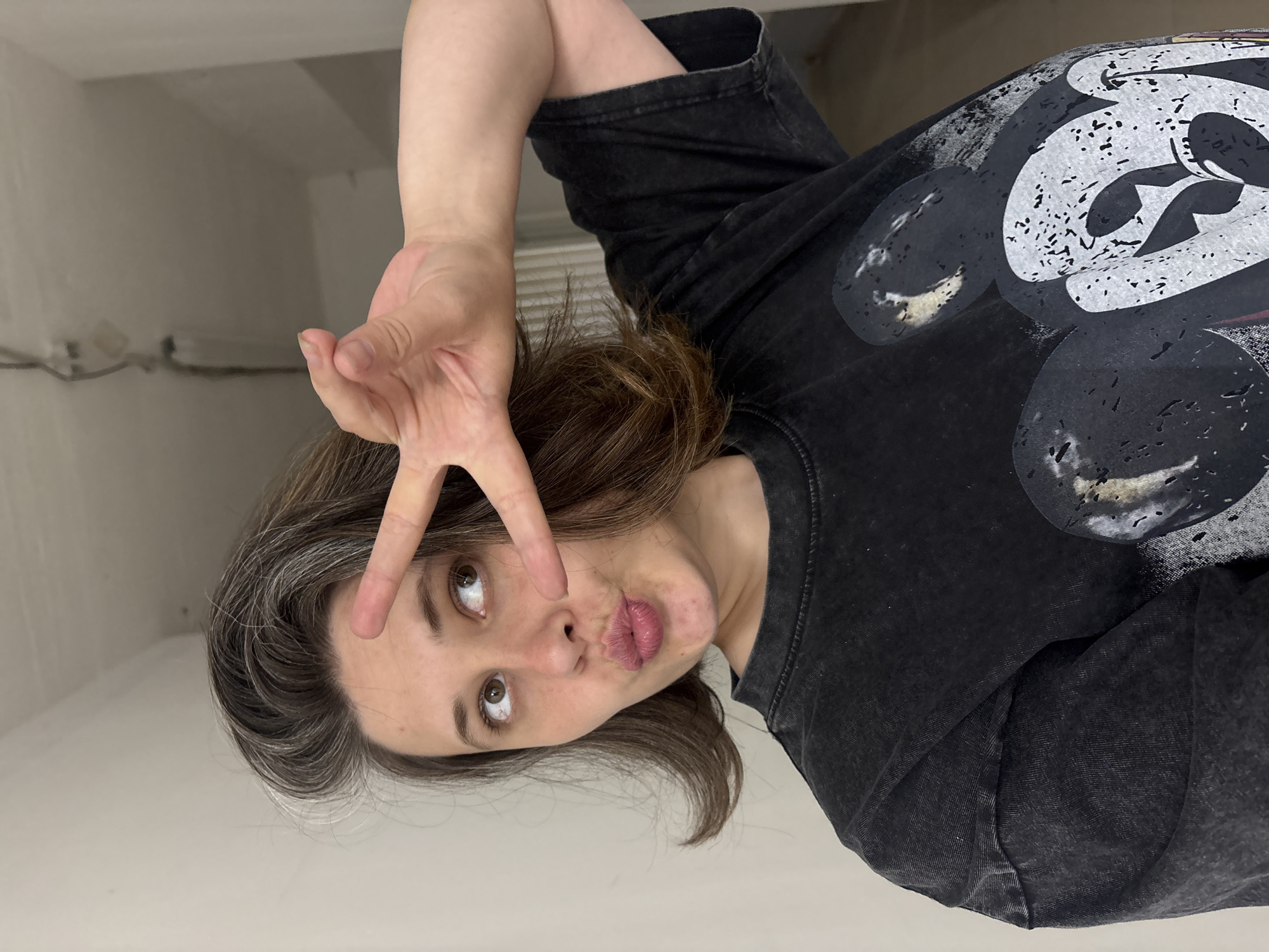 A young woman with brown hair, streaked with gray, is pictured from the chest up. She is wearing a black t-shirt with a large graphic of Mickey Mouse's face on it. She is making a peace sign with her right hand, holding it up to her forehead. Her lips are pursed in a playful expression. Her eyes are open and looking slightly to her left. The background is a plain, light-colored wall. The image is oriented vertically, with the top of her head towards the left side of the frame. The lighting is bright and even, suggesting an indoor setting with artificial light. There is no visible text in the image. The mood of the image is casual and fun.