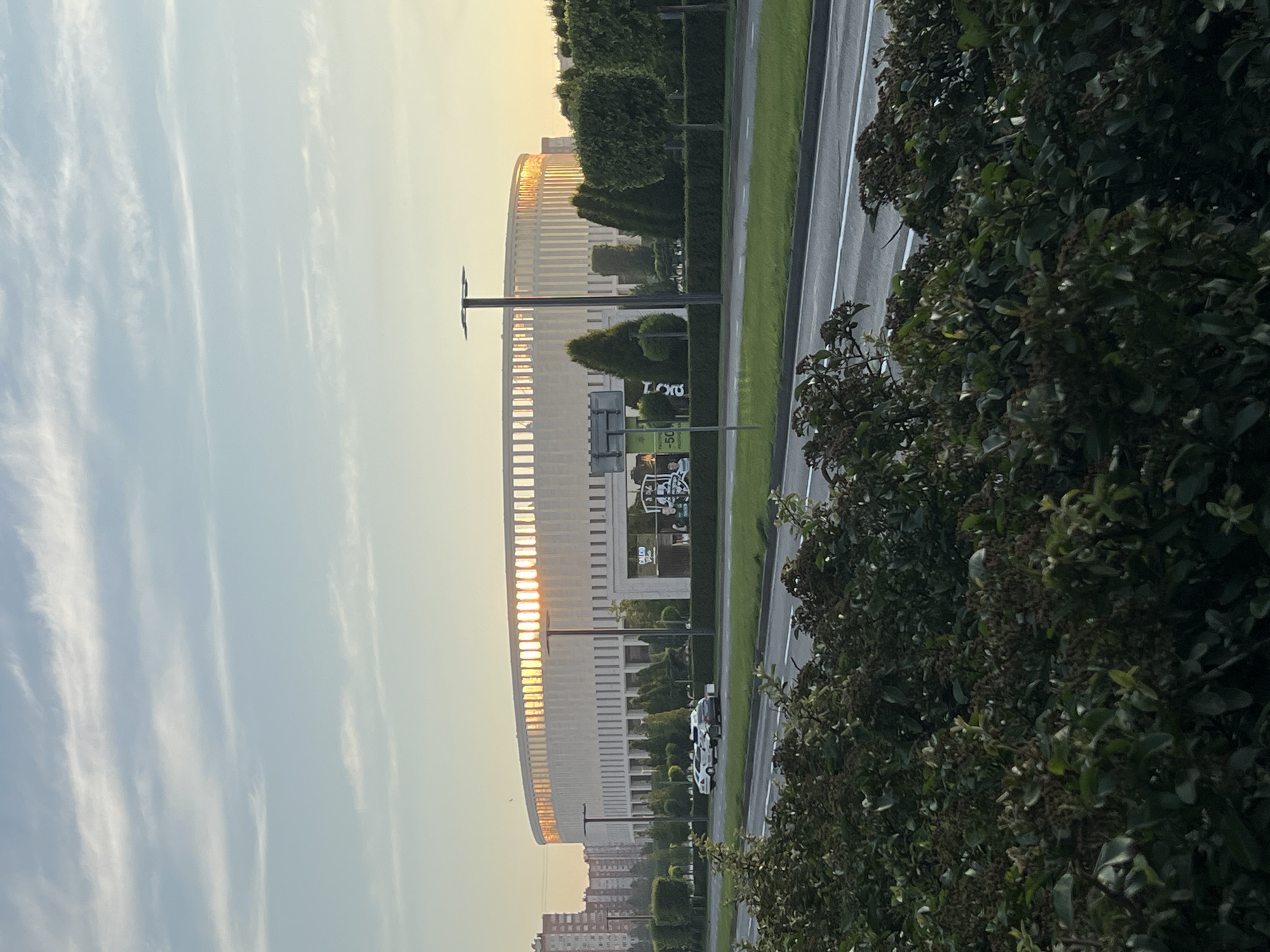 In Krasnodar, Russia, the image captures a wide-angle view of a modern stadium, likely the FC Krasnodar Stadium, during a sunset. The stadium's distinctive circular design is illuminated by the golden rays of the setting sun, which peek through the vertical elements of its facade, creating a warm glow. The stadium is situated in a well-maintained urban environment, with a broad, paved road in the foreground, bordered by a neatly trimmed green lawn and mature trees. Several cars are parked along the road, and building structures are visible in the background. The sky is a soft blue, with wispy clouds, indicating pleasant weather. The overall scene conveys a sense of tranquility and architectural impressiveness at dusk.