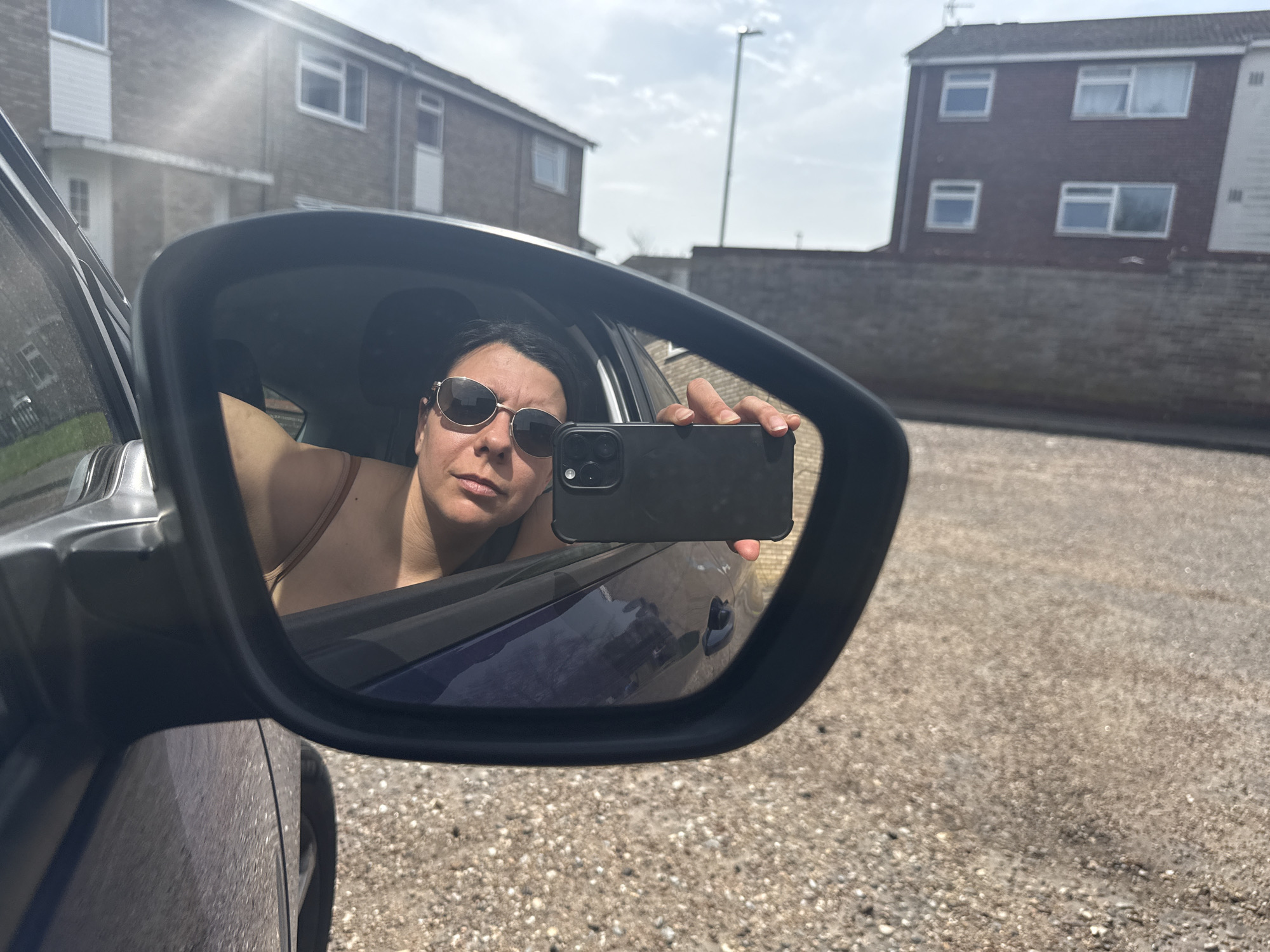 A woman with dark hair and sunglasses is taking a selfie in the side mirror of a blue car. She is holding a black smartphone in her right hand. The car appears to be parked on a paved road with pebble textures. In the background, there are two brick buildings with windows and a light post, indicating a residential area of Gorleston-on-Sea, United Kingdom. The sky is bright with scattered clouds and the sun is causing glare on the left.