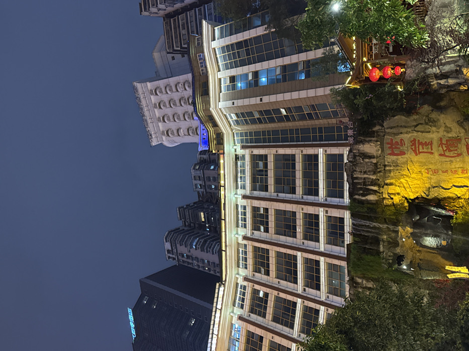 The image displays an illuminated urban architectural complex at night in Yuzhong District, Chongqing, China. The upper portion of the frame shows a dark, overcast night sky devoid of stars.
In the background, multiple high-rise buildings are visible; a dark skyscraper with blue neon signage on its rooftop stands to the far left, while a lighter-colored building with multiple levels of recessed, rounded windows is prominent to the right.
The central foreground features a large, multi-story building with a curved, tiered facade and numerous grid-patterned, reflective windows. This structure is outlined and highlighted by horizontal white light strips along its upper levels. A small sign with the Chinese characters "观景台" (Guānjǐng tái - Observation Deck) is visible on a lower section of this building.
Directly beneath this structure, a prominent, rugged rock face is heavily illuminated in yellow and orange light. A narrow waterfall cascades down from the edge of the building above onto this rock face. Green vegetation, consistent with moss or small plants, adheres to sections of the rock near the waterfall. Large red Chinese characters, reading "洪崖洞" (Hongyadong) followed by smaller characters "民俗风貌区" (Mínsú fēngmào qū - Folk Culture Street/Area), are painted or carved into the glowing rock face.
To the lower right, partially obscured by dark green foliage, is a traditional Chinese-style pavilion with a dark, layered tiled roof and visible wooden pillars. Three red, circular lanterns hang from this pavilion structure. No individuals or discernible human activity are present within the scene.