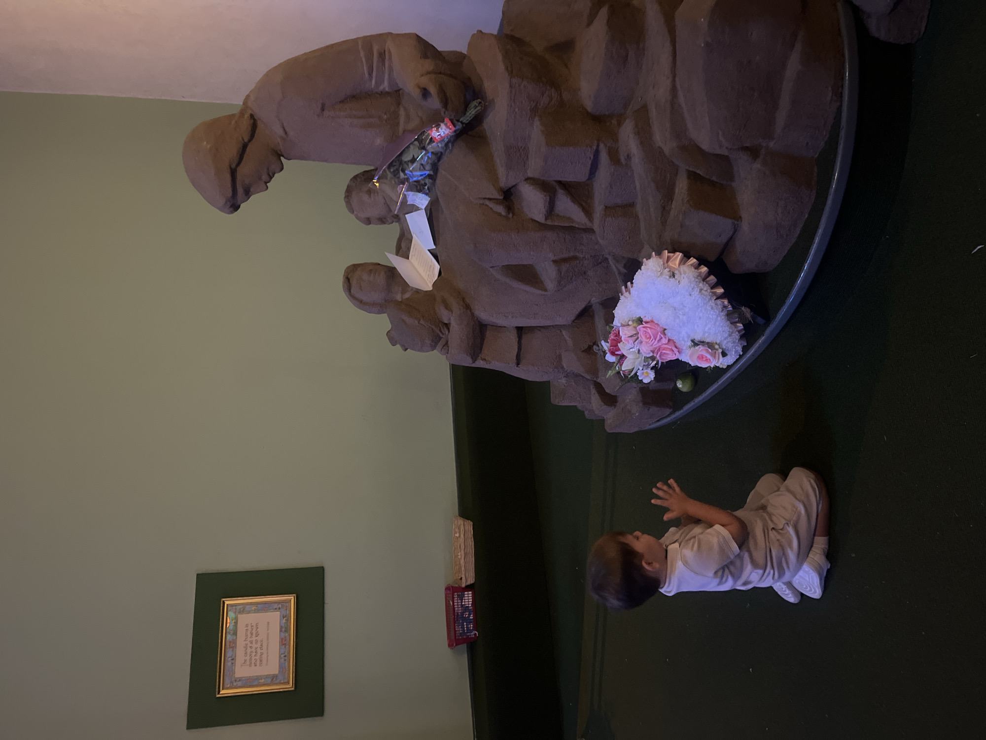 A toddler kneels on a dark green carpet in front of a large, brown, stone sculpture. The sculpture depicts a robed figure appearing to read from a piece of paper to three smaller figures. A bouquet of pink and white flowers rests at the base of the sculpture. The toddler is wearing a light-colored outfit and white sneakers, and has their hands clasped in front of them. In the background, a light green wall is visible, with a framed picture hanging on it. Two wicker baskets are on a dark surface behind the toddler. This scene is located inside a building in Liverpool, United Kingdom.