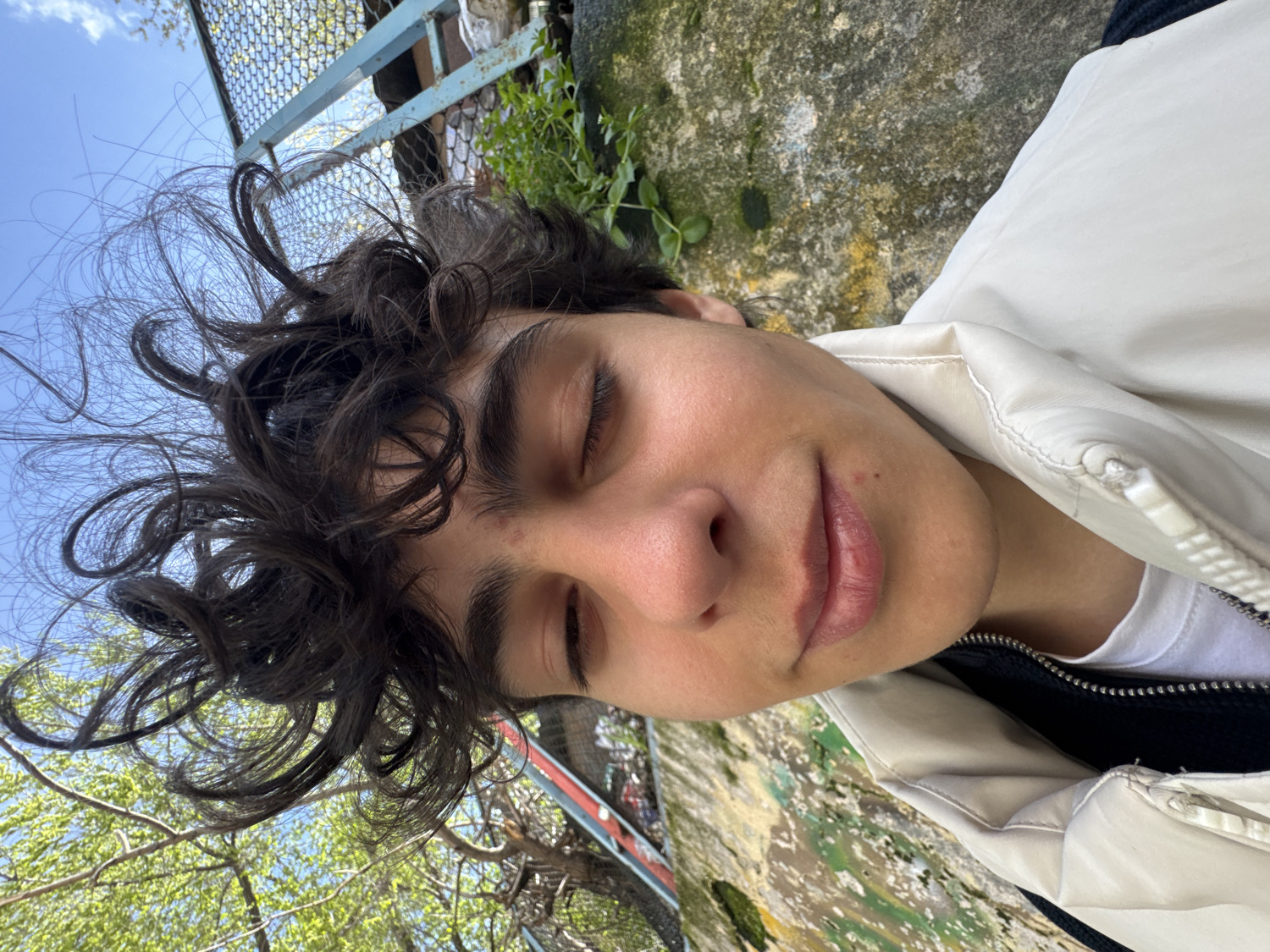 The image presents a close-up, outdoor portrait of a young person, depicted from the chest up with their head tilted and eyes closed. The person has dark, voluminous curly hair and a relaxed, slight smile on their lips, conveying a sense of serenity or enjoyment of the moment. They are wearing a light-colored, possibly off-white or cream, zippered jacket over a white t-shirt, with a dark, zippered layer underneath.
The setting is outdoors on a bright, sunny day, suggested by the clear blue sky visible in the upper left of the frame. In the background, behind the person's head, there is a blue metal fence or grid structure. To the right and bottom, a textured, weathered surface, possibly concrete or stone, is visible, featuring patches of green moss and other discoloration. Sparse green foliage and tree branches with fresh leaves are also present, indicating a natural or semi-natural environment within an urban or semi-urban context. The clear lighting signifies daylight. The scene is located in Yerevan, Armenia. No visible text is present in the image.