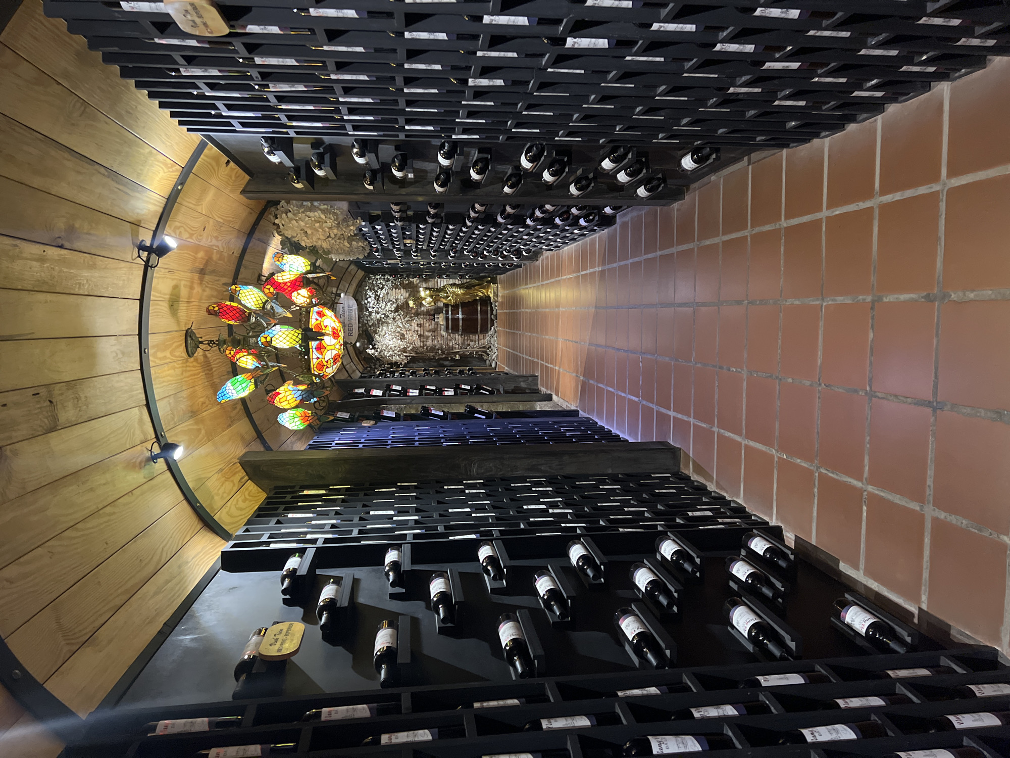 This is a long shot of a wine cellar, possibly in Da Lat, Vietnam, featuring rows of dark wine racks filled with bottles. The ceiling is made of wooden planks, and a chandelier with colorful stained-glass parrot figures hangs in the center, illuminating the space. The floor is tiled with reddish-brown square tiles. In the background, there is a golden statue of a woman next to a wooden barrel, and decorative elements like a stone wall and white floral arrangements. The lighting is dim, creating a warm and inviting atmosphere characteristic of a wine cellar.
