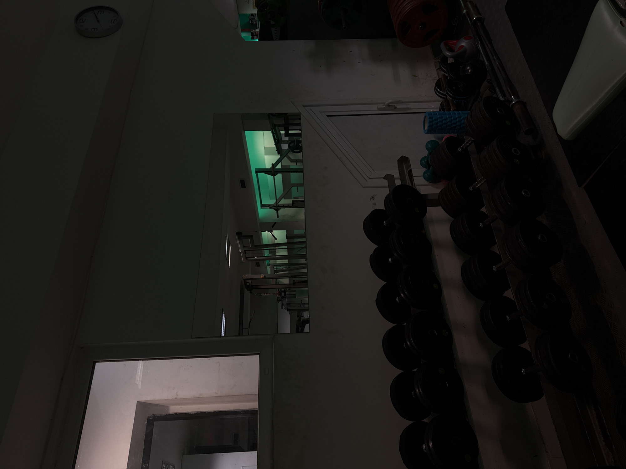 The image depicts the interior of a gym, likely in Varna, Bulgaria, based on the prompt. A clock is visible in the top-left corner of the frame. Through an open doorway and a window-like opening above, the space appears to contain weightlifting equipment, illuminated by green lights. Dumbbells are arranged on a rack on the right-hand side of the frame. Other gym equipment can be seen in the background. The image is dimly lit.