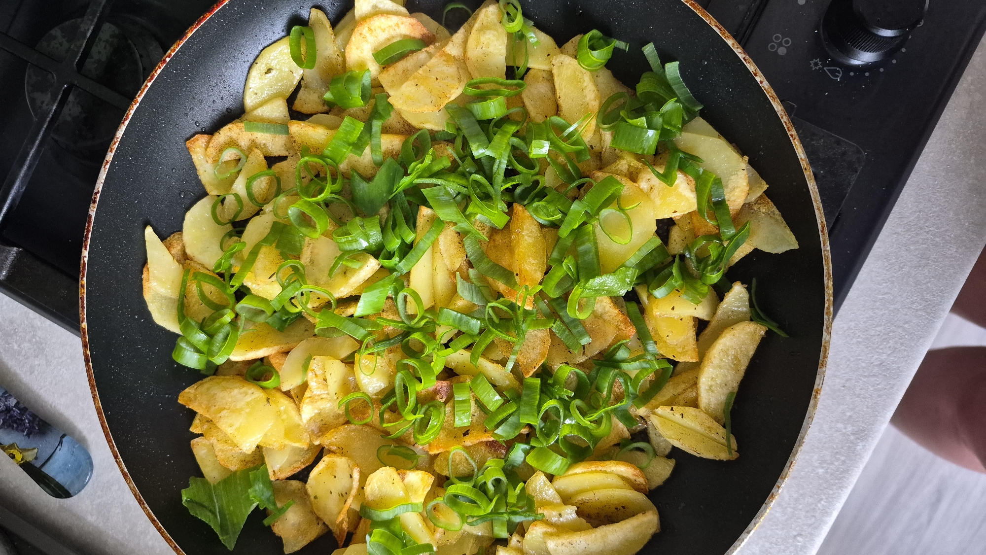 A close-up overhead shot shows a black frying pan filled with golden-brown fried potato wedges and sliced green onions. The potatoes appear seasoned with herbs and spices, and some have slightly crispy edges. The green onions are thinly sliced and scattered generously over the potatoes, providing a vibrant green contrast. The pan is placed on a stovetop, with a glimpse of a burner and control knobs visible in the lower right corner. The background is out of focus, suggesting an indoor kitchen environment. The image does not provide any information about the city of Krauja, Latvia.