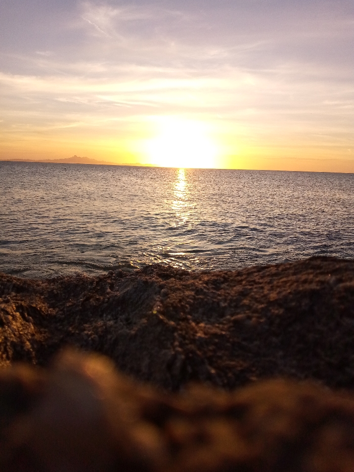 The image presents a panoramic view of a sunset over a body of water, observed from the coast of Juan Griego, Venezuela, looking westward over the Caribbean Sea. A brilliant, white-hot sun is positioned centrally on the horizon line, casting an intense, shimmering path of light across the water's surface towards the viewer. The sky transitions from a pale lavender at the zenith to vivid orange and yellow hues near the horizon, with subtle, wispy cloud formations overhead. On the distant left horizon, a dark, undulating landmass is silhouetted, showing irregular topography, consistent with geographical features visible from Juan Griego. The immediate foreground, occupying approximately the lower third of the frame, consists of dark, heavily textured, out-of-focus rock formations or coarse sediment, indicating a low-angle photographic perspective near the water's edge. No human figures, vessels, or specific activities are present within the frame.