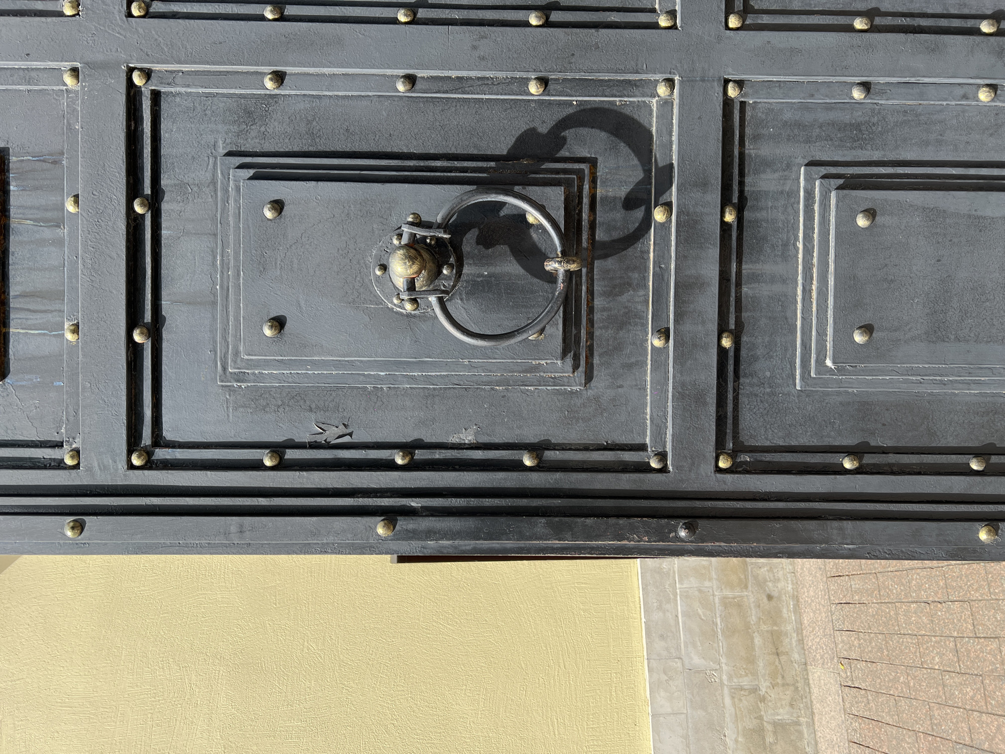 The image shows a portion of a large, dark gray, possibly metal door with decorative brass studs and a central metal ring handle. The door has a panelled design with rectangular frames. The handle casts a distinct shadow. The bottom edge of the door is visible, revealing a yellow wall and a tiled pavement. The location is unknown.