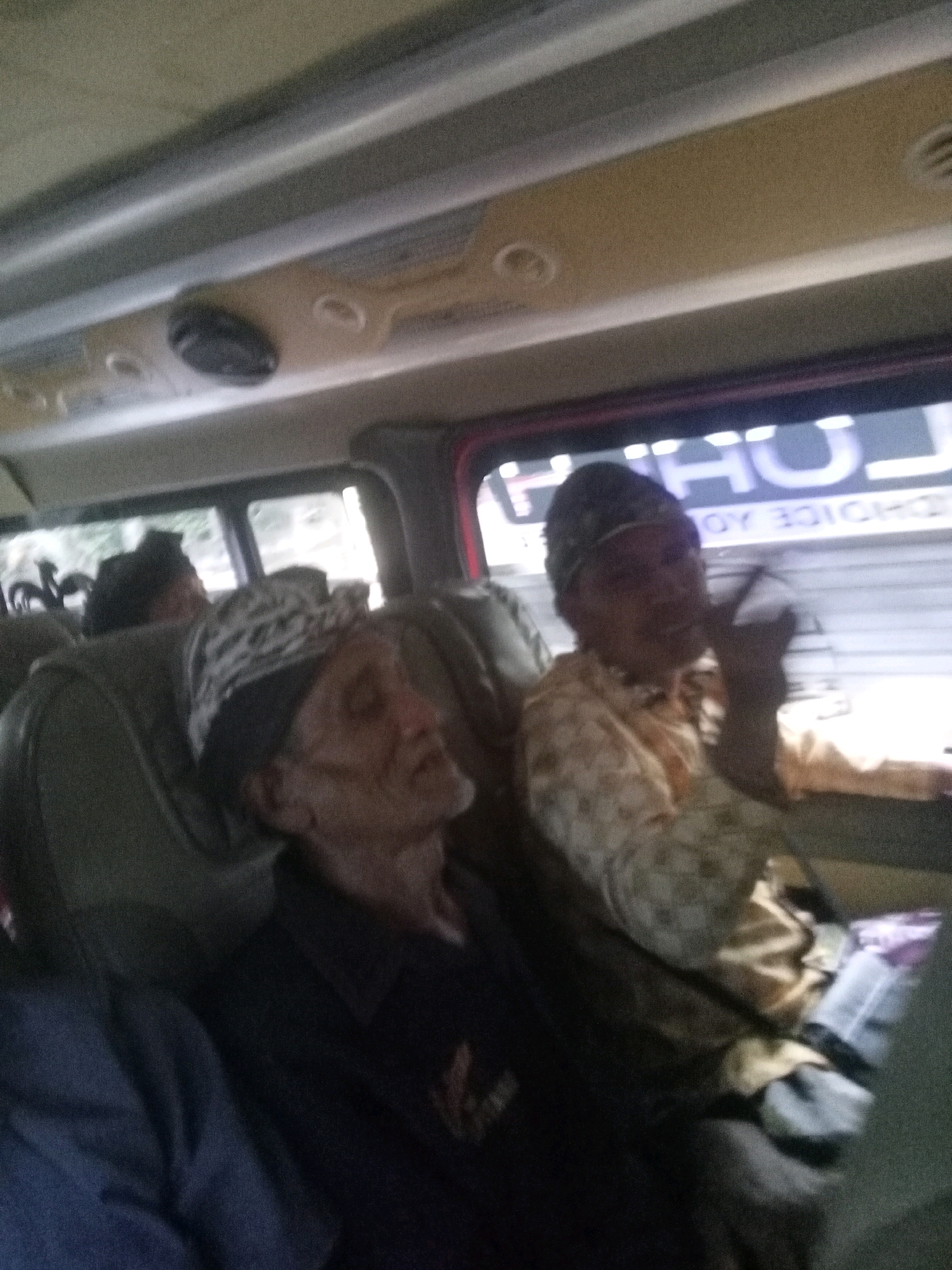 This is a blurry, low-light, medium-shot photograph taken from inside a vehicle, possibly a van or bus, in Pekutatan, Indonesia. The image is dominated by the interior of the vehicle, including the ceiling with overhead compartments and lights, and the seats occupied by several individuals.
In the foreground, a person wearing a patterned head covering and a dark shirt is seated, with their head tilted back and eyes closed, suggesting they are asleep or resting. To their right, another individual is seated and appears to be awake. This person is wearing a light-colored, patterned shirt. The windows of the vehicle are visible, showing a blurred outdoor scene. One window has text superimposed on it, which appears to be in a foreign language, possibly with characters like "U," "H," and "OY."
The overall impression is that of passengers traveling within a vehicle, likely on a journey, in a semi-urban or rural setting given the mention of Pekutatan, Indonesia. The lighting is dim, contributing to the blurriness and lack of sharp detail in the image.