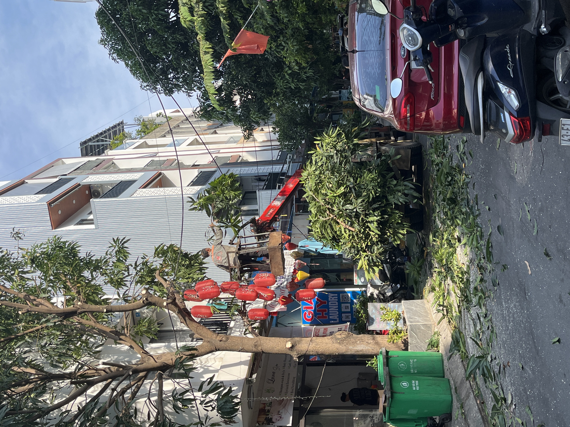 This image captures a bright, sunny daytime street scene in Da Nang, Vietnam, showing a mix of urban activity and ongoing maintenance. The primary activity depicted is tree pruning; a worker is elevated in a red crane or lift truck, actively trimming branches from a large tree. Freshly cut leaves and small branches are scattered across the pavement and sidewalk, indicating recent work.
The street is lined with multi-story buildings featuring modern architectural elements, some with balconies and visible air conditioning units. On one of the rooftops, what appears to be solar panels are installed. Parked vehicles include a red Ford Focus car and a dark Yamaha Grande scooter, with another scooter partially visible, typical of Vietnamese streetscapes.
Numerous objects add detail to the scene. Several red, traditional-style lanterns hang from the branches of a tree near a shopfront. Two green public trash bins are visible on the sidewalk, bearing the Vietnamese text "HÃY BỎ RÁC VÀO THÙNG" (Please put trash in the bin). A small red flag, possibly the Vietnamese flag, is attached to a pole in the background.
Visible text from various signs includes "Luna Spa" offering services like "Shampoo-wash & Style," "Body Massage," and "Eyelash Extensions." Another sign advertises "GIA HY LAUNDRY." Partial text like "JAHY TRAVEL CAR" is also discernible. The license plate fragment "L3-F1" is seen on one of the scooters. The clear blue sky and bright sunlight suggest good weather conditions.