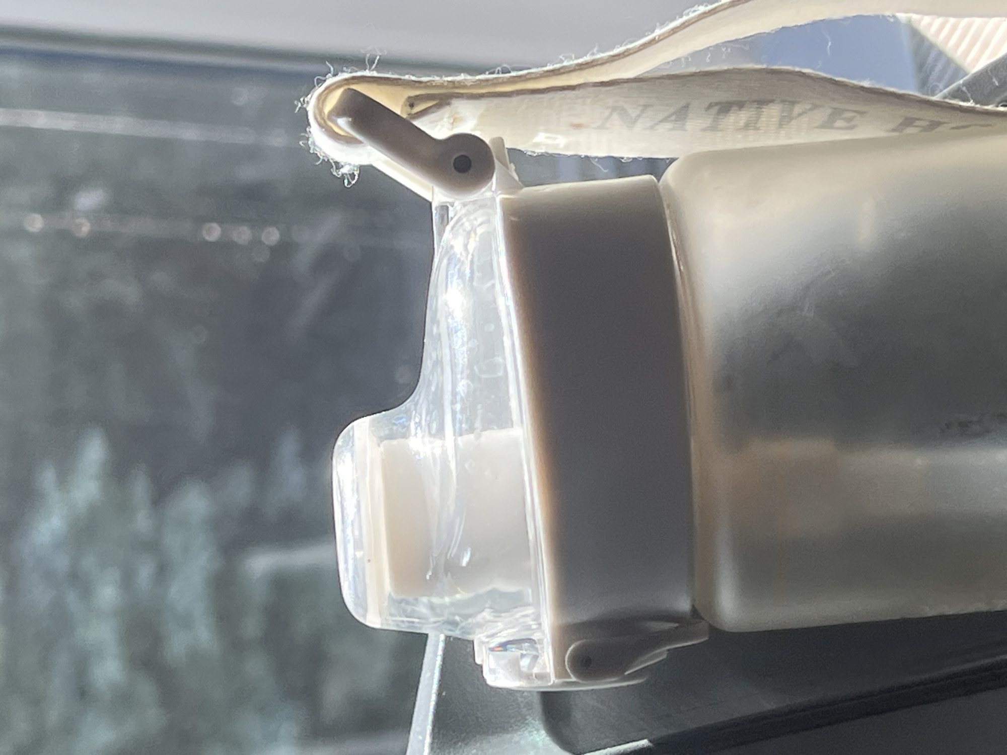 A close-up, slightly angled shot focuses on the translucent lid and upper portion of a beige water bottle. The lid is clear plastic with a white dispensing mechanism. A beige fabric strap, with the faint visible lettering "NATIVE F", is attached to the lid via a grey clip. The bottle itself appears to be made of frosted plastic. In the blurred background, the out-of-focus shapes of green foliage are visible, suggesting an outdoor setting, possibly near trees. Sunlight is casting bright highlights on the bottle and strap, and creating a bokeh effect of light circles in the background. The scene does not provide any specific visual cues to pinpoint a location in Moscow, Russia.