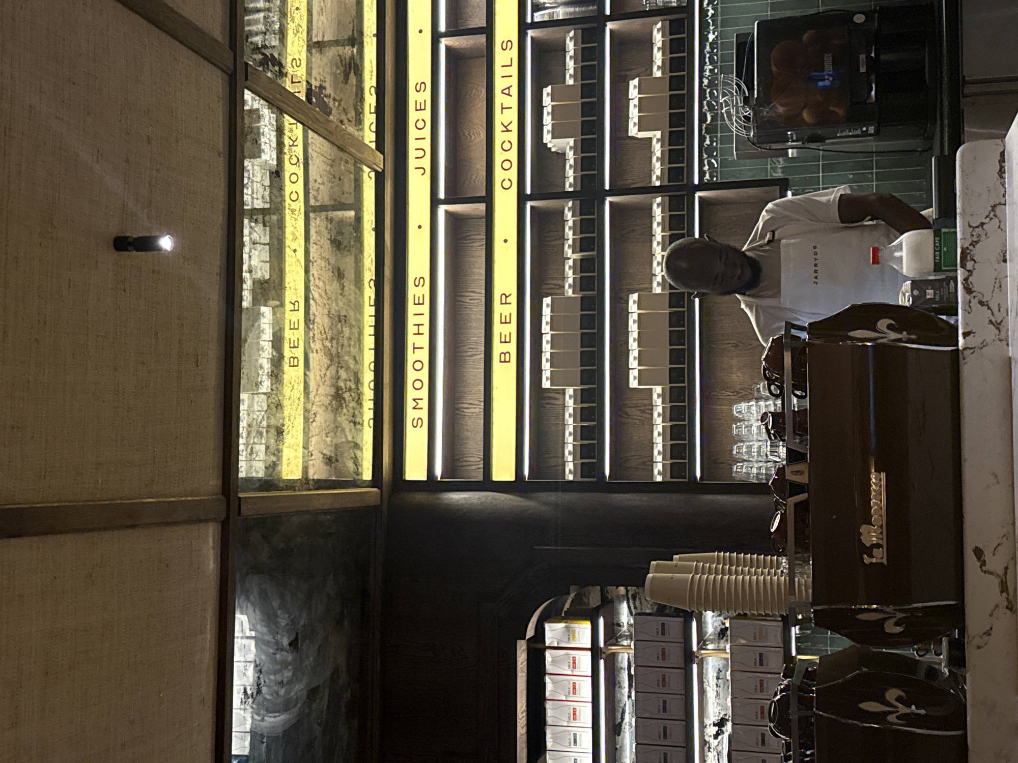 The image depicts the interior of a JARRYDS restaurant, located in Cape Town, South Africa. The setting is a bar area. Behind the bar, there are vertical displays with the words "BEER," "SMOOTHIES," "JUICES," and "COCKTAILS" written on them. There is also what appears to be a menu displayed behind the glass, with other menu items listed on it. Stacks of neatly arranged product packaging are visible below the counter.
A person wearing a white shirt and an apron with the name "JARRYDS" on it is working behind the bar. There is also an orange juice machine on the counter next to the person. An espresso machine is also visible. Stacks of paper cups are placed on the counter.