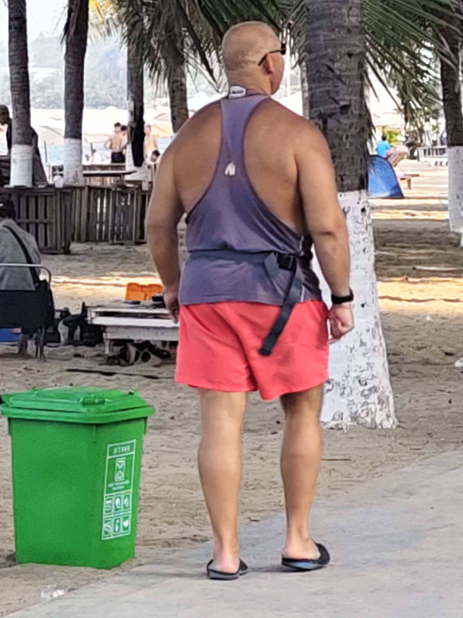 A bald man wearing sunglasses, a purple tank top, and red shorts stands on a sandy pathway in front of a palm tree. He is facing away from the camera. To his left is a green trash can with symbols on its side. In the background, palm trees and people are visible near tents and beach structures. The scene appears to be a public beach area in Nha Trang Ward, Vietnam.
