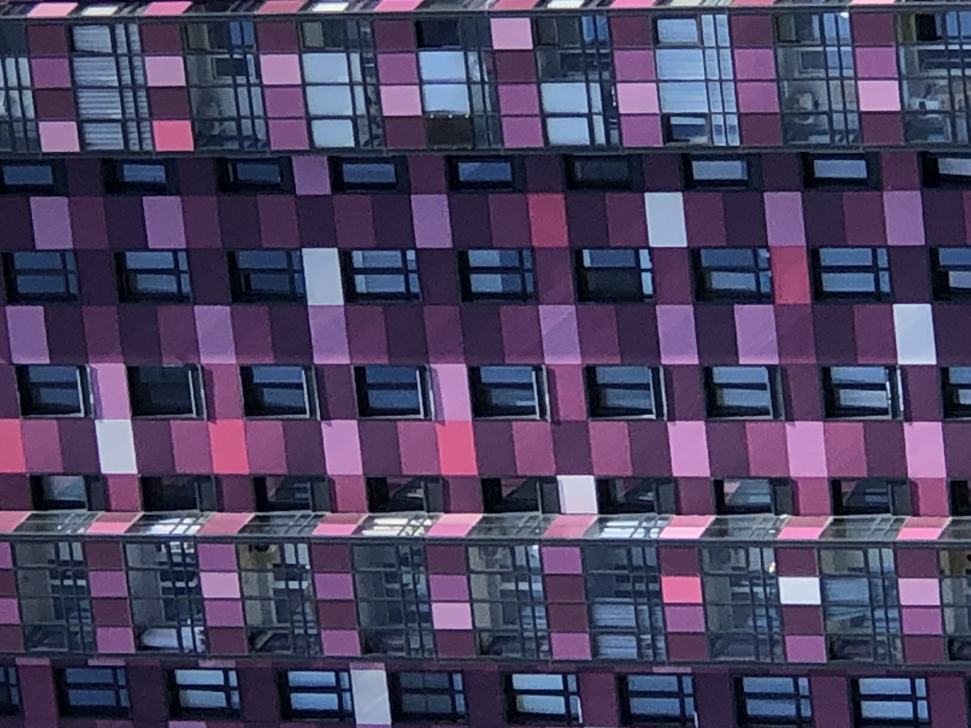 The image shows a close-up of a modern apartment building facade in Saint Petersburg, Russia. The building features a striking, checkered design with alternating square panels of various shades of purple and pink. The panels are interspersed with numerous windows, some of which are dark and reflective, suggesting they are glass. Several balconies are visible, enclosed with glass and dark metal framing. The architectural style appears contemporary, characterized by geometric patterns and a vibrant color palette. The lighting suggests it is daytime, with sunlight illuminating parts of the facade and casting subtle shadows. No people are directly visible in the image, and the focus is solely on the building's exterior.