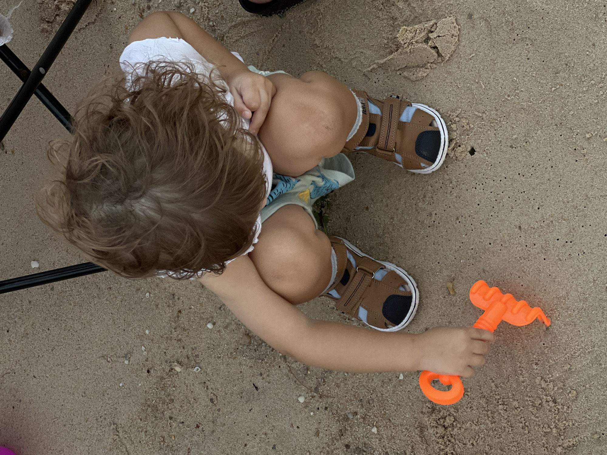 The overhead shot shows a young child sitting on sandy ground. The child has light brown hair and is wearing a white top and what appears to be a printed fabric underneath. Their legs are tucked up, and they are wearing brown sandals with blue accents. The child is holding an orange toy rake. The scene is outdoors, as indicated by the sand and natural lighting. The time of day and weather are not discernible. The background is mostly sand with some small shells scattered about, and a portion of a black chair is visible in the upper left corner. The overall impression is of a child playing on a beach or in a sandy area.