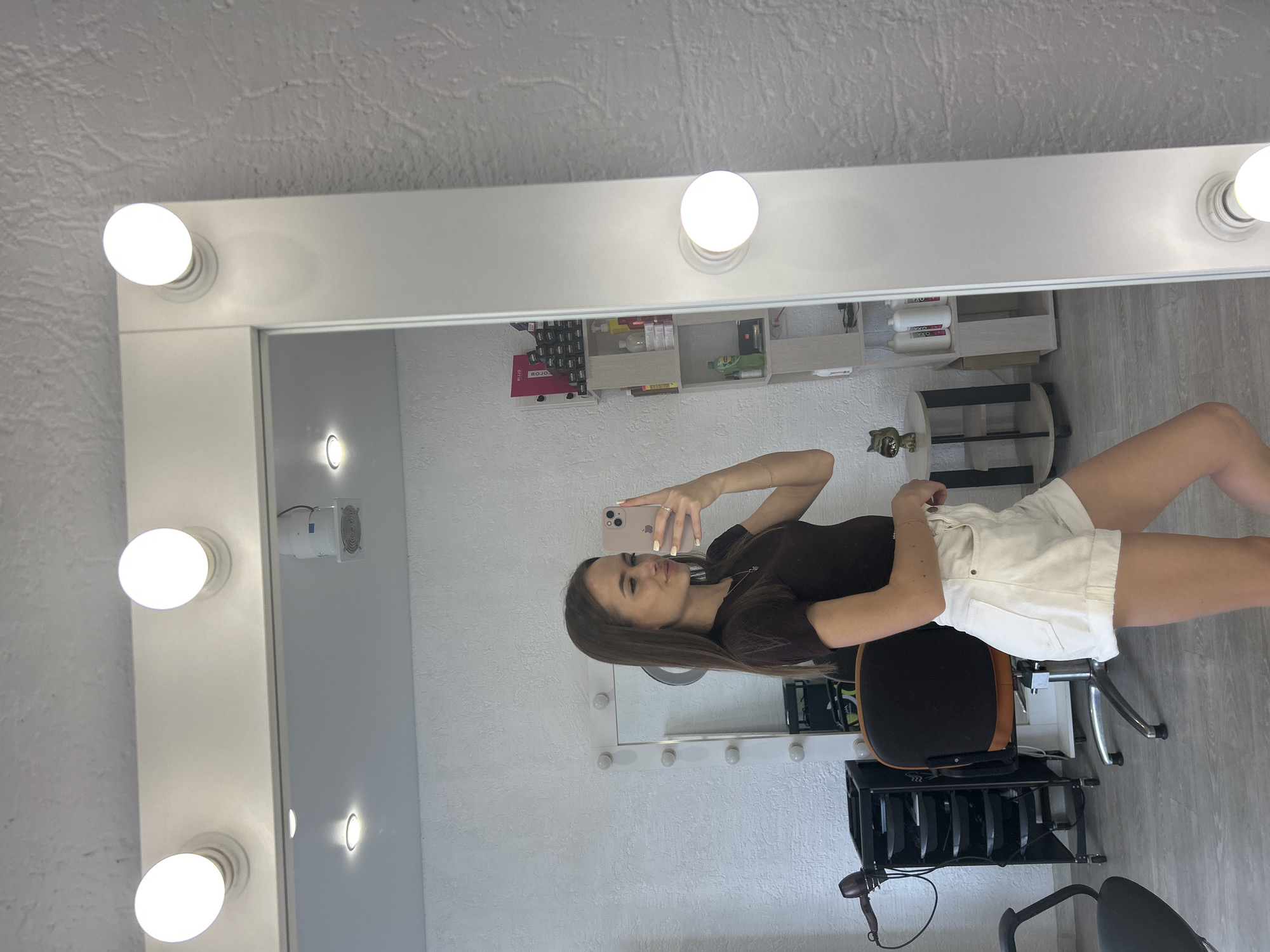 A young woman with long brown hair, wearing a dark brown t-shirt and white shorts, stands in front of a large illuminated mirror, taking a selfie with her phone. The mirror is framed by light bulbs. In the background, a hair salon or similar establishment is visible, with shelves stocked with bottles, a hairdryer on a stand, and chairs. The walls have a textured finish.