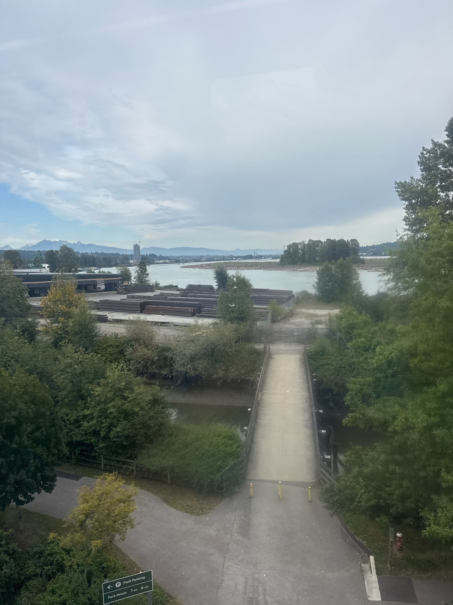 This image, captured from a high vantage point, likely a building window, overlooks a section of the Fraser River in New Westminster, Canada. The scene is dominated by a wide, calm river under an overcast sky, with distant mountains forming a hazy backdrop. In the foreground, a concrete pedestrian bridge spans a narrow waterway or inlet. The bridge's path leads towards a paved area with some bollards and a fire hydrant.
To the left of the bridge, along the riverbank, is an industrial area with stacks of metal beams and what appear to be large trucks. Trees and lush green foliage line the riverbanks and surround the bridge and industrial site, suggesting a mix of urban infrastructure and natural elements. A signpost indicating "Park Parking" and "Park Hours 7 am - 8 pm" is visible in the lower left corner, suggesting a park area is nearby. The overall impression is one of a working riverfront, where industry coexists with recreational spaces.