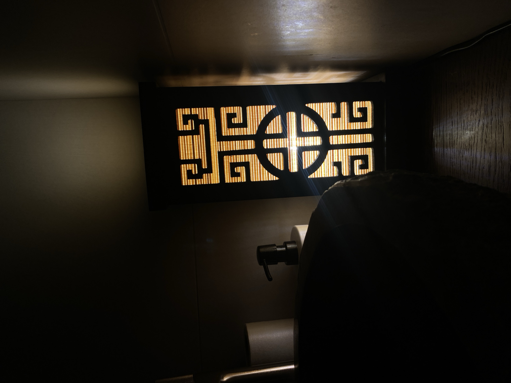 The image shows a dark, dimly lit bathroom interior. A decorative, rectangular light fixture with a black frame and a geometric pattern is mounted on the wall. The pattern consists of horizontal bars and stylized spiral shapes surrounding a central circular design with a cross. A light source shines through the pattern. Beneath the light fixture, a toilet paper dispenser and a toilet are visible. A portion of a person's body is in the foreground, obscuring some of the scene. The wall appears to be a light color. The location is unknown and cannot be determined based on the image.