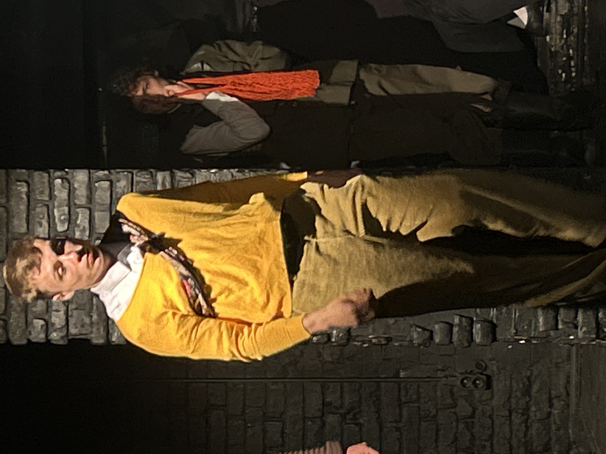 The image depicts two individuals lying on what appears to be tiered platforms or levels constructed of dark material resembling brick or stone. The setting is ambiguous, but seems to be a stage or theatrical setting, possibly in a city of Unknown, Unknown.
The person in the foreground is wearing a yellow sweater over a white collared shirt and tan trousers. They are lying on their side, facing the viewer, and appear to be of light complexion.
Above them, another individual is lying on a similar platform. They are wearing darker clothing and an orange scarf or neckwear. Their pose seems similar to the person below.
The backdrop is dark and the lighting accentuates the individuals on the platforms. The precise context or narrative of the scene is not immediately discernible.