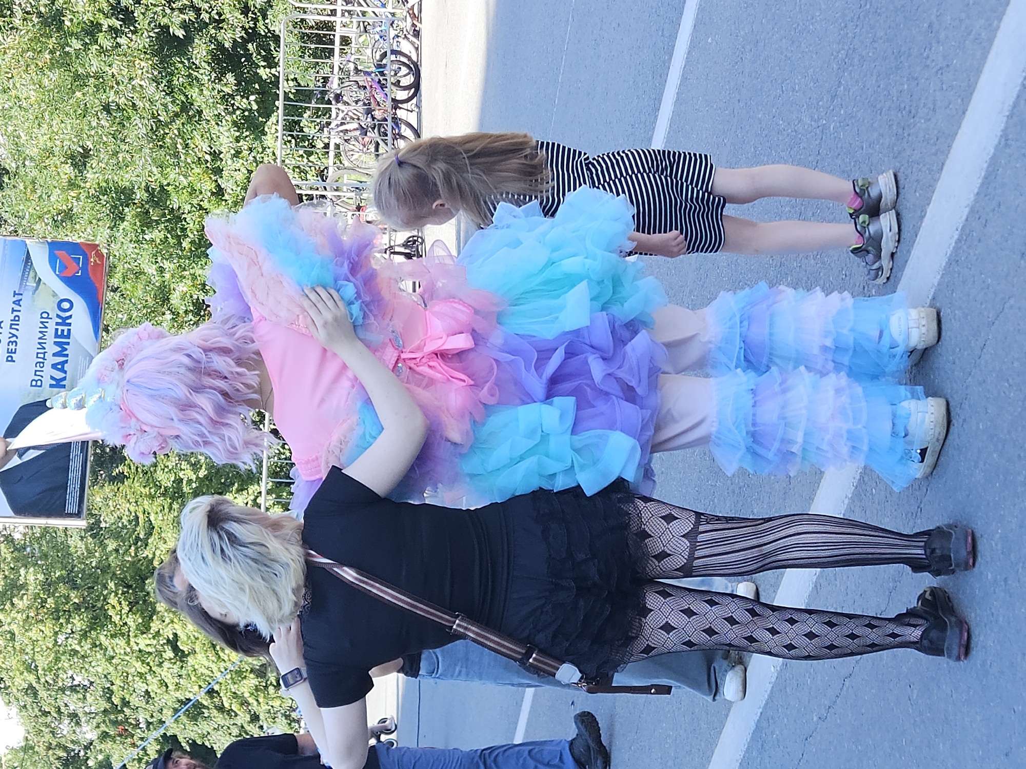 The image shows three individuals standing on a paved surface, likely a street or sidewalk, in an outdoor urban environment. In the foreground, from left to right, is a person wearing black ruffled shorts, patterned black stockings, and black sneakers. They have blonde hair and are facing away from the camera, with their arms around another person. The second person is in the center, wearing a colorful, layered outfit with pastel pink, blue, and purple ruffles, and a unicorn-like headdress. They are also facing away from the camera, with their arms around the first person. To the right of these two is a young girl in a black and white striped dress and sneakers, facing away from the camera.
Behind the individuals, a white metal fence is visible, with bicycles parked behind it. In the background, there are trees and a large banner with images and text in Russian. The text on the banner appears to be political, with the name "Владимир КАMEKO" prominent. The overall scene suggests a public event or gathering. The presence of the banner and the style of dress of the central figure hint at a festive or celebratory occasion, possibly a parade or festival.
The location is identified as Ulyanovsk, Russia. The paved ground, urban infrastructure like the fence and bicycles, and the type of trees visible are consistent with an urban setting in Russia. The banner suggests a political campaign or event taking place in Ulyanovsk. The lighting indicates it is daytime, likely sunny.