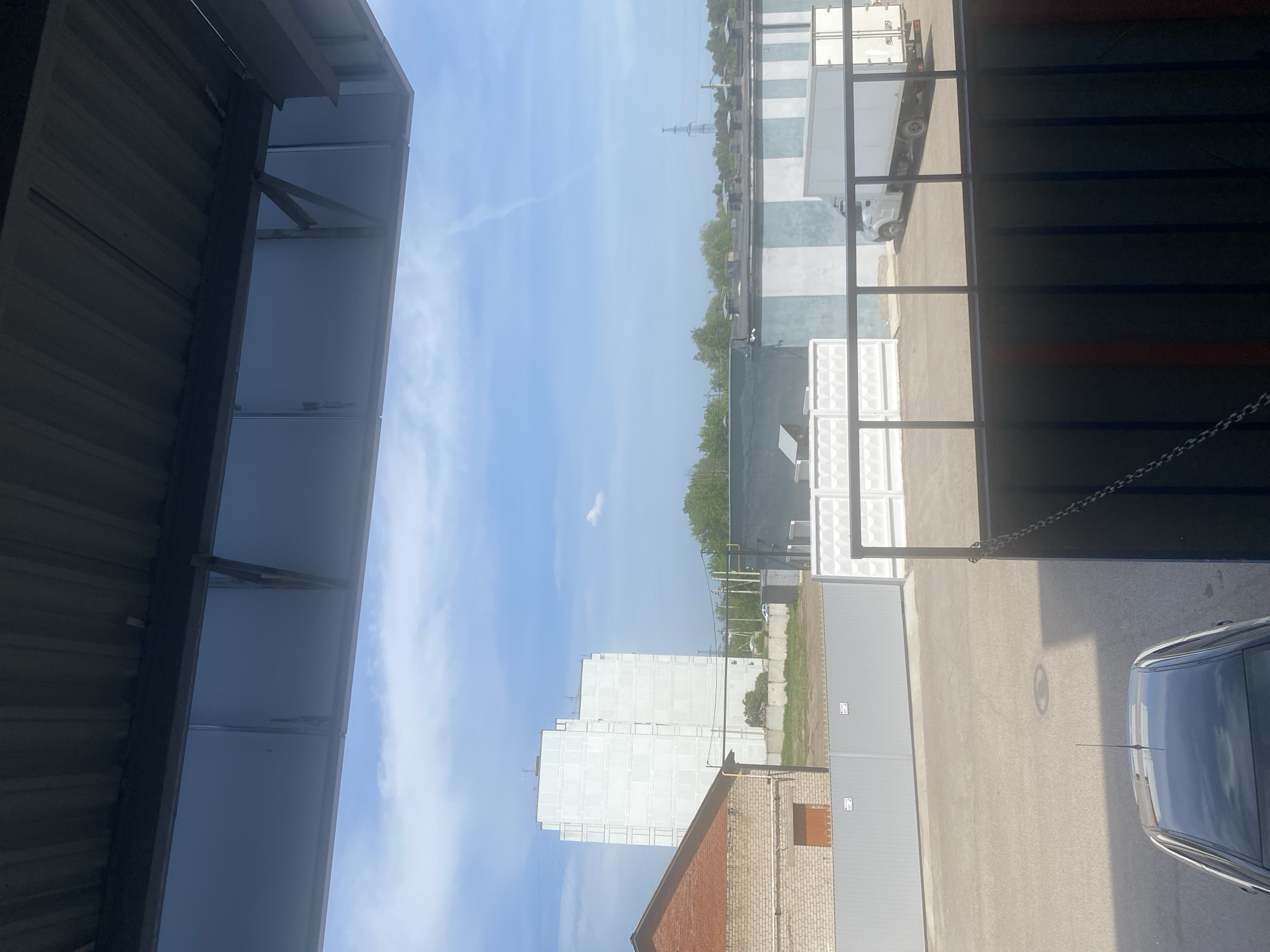 The image captures an outdoor scene in what appears to be an industrial area, potentially located in or near the city of Chaikovsky, Russia. The perspective is taken from underneath a partially visible metallic structure, possibly a roof or awning, with steel supports and metallic paneling. A blue sky with light clouds is visible in the background.
Across a paved area, there are several large, white containers or modular units, and a few parked trucks. Beyond the containers, there is a line of trees and several buildings, including a tall, white apartment building that dominates the skyline. A brick building is visible near the bottom of the image with a small orange vent on the side. A car is seen at the right edge of the frame. There is no indication of people in the image. An electricity pylon is noticeable in the distance.