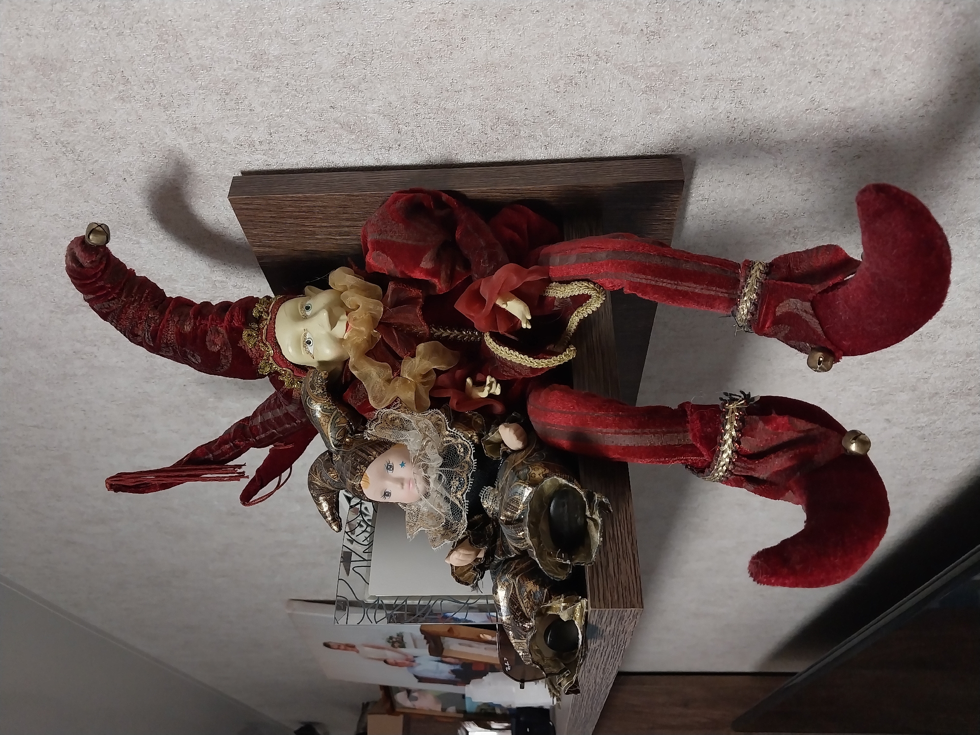 The image shows a shelf mounted on a textured, light-colored wall. On the shelf are two jester dolls. The doll on the right is larger and sits upright, wearing a red and gold jester costume with a tall, pointed red hat adorned with red fringe and a small bell. The doll has a pale face with wide eyes and a smiling expression. Its legs are long and red with gold trim and red shoes with bells. The doll on the left is smaller and appears to be a baby doll dressed in a jester costume of gold and dark patterned fabric, with a ruffled collar and a smaller jester hat. Behind the dolls, on the shelf, are framed pictures, including one with two people. The shelf itself is made of wood and is positioned in an indoor setting, likely a home. The lighting suggests ambient room lighting. There is no indication of the specific location within Dnipro, Ukraine.