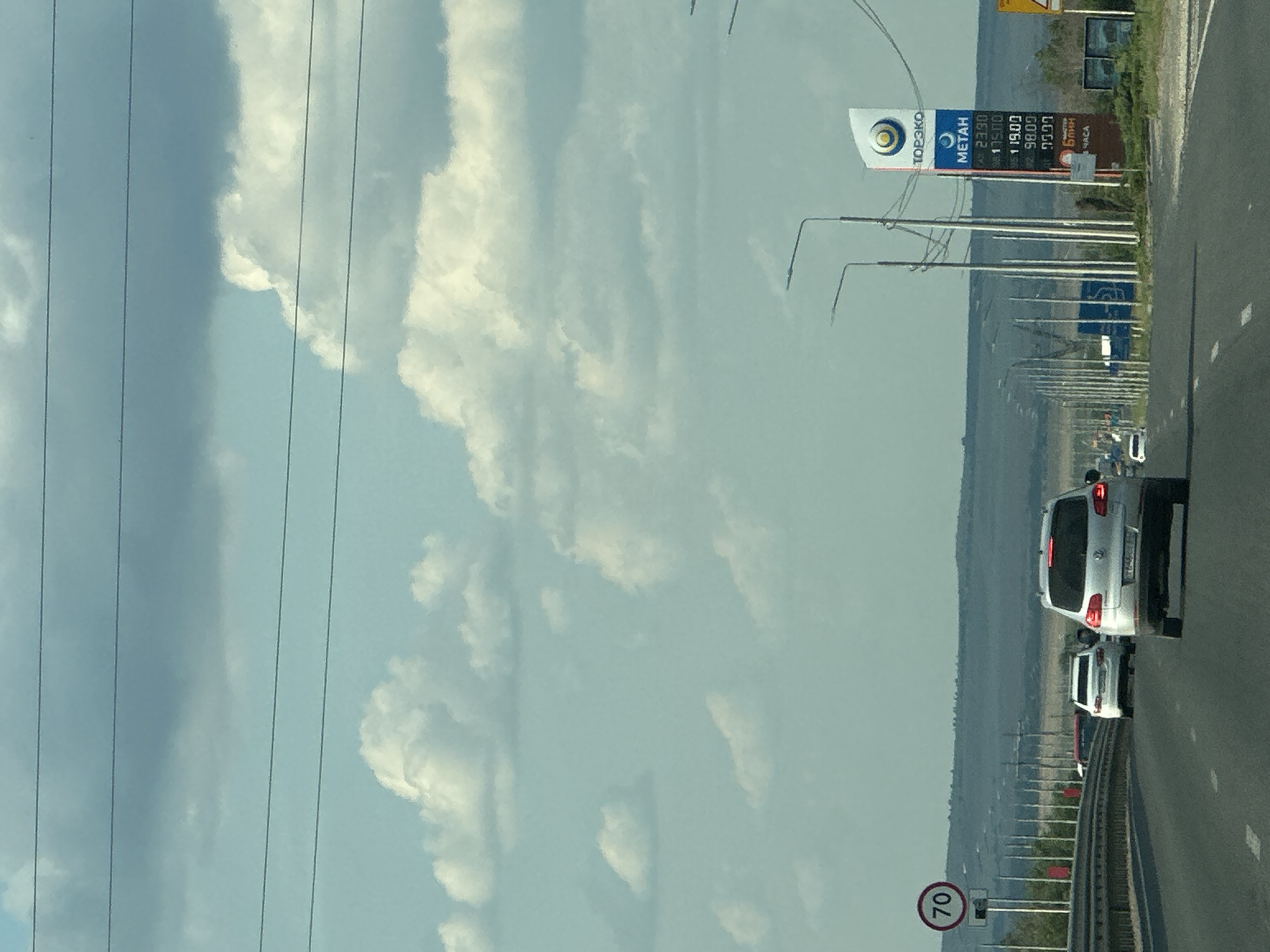 The image shows a highway scene in Zorinskii, Russia. The view is from a vehicle traveling on a multi-lane asphalt road. On the right side of the road, there is a gas station with a prominent sign displaying fuel prices, including "METAH" priced at 23.90, 19.00, 98.00, and 29.97. Several streetlights line the right side of the highway. On the left side of the road, there is a speed limit sign indicating "70". Several cars are visible on the road, including a silver SUV in the foreground, a white sedan behind it, and other vehicles further down the highway. The sky is partly cloudy with a light blue hue, and the landscape in the distance appears dry and hilly. The overall impression is a typical highway scene in a rural or suburban setting in Russia.
