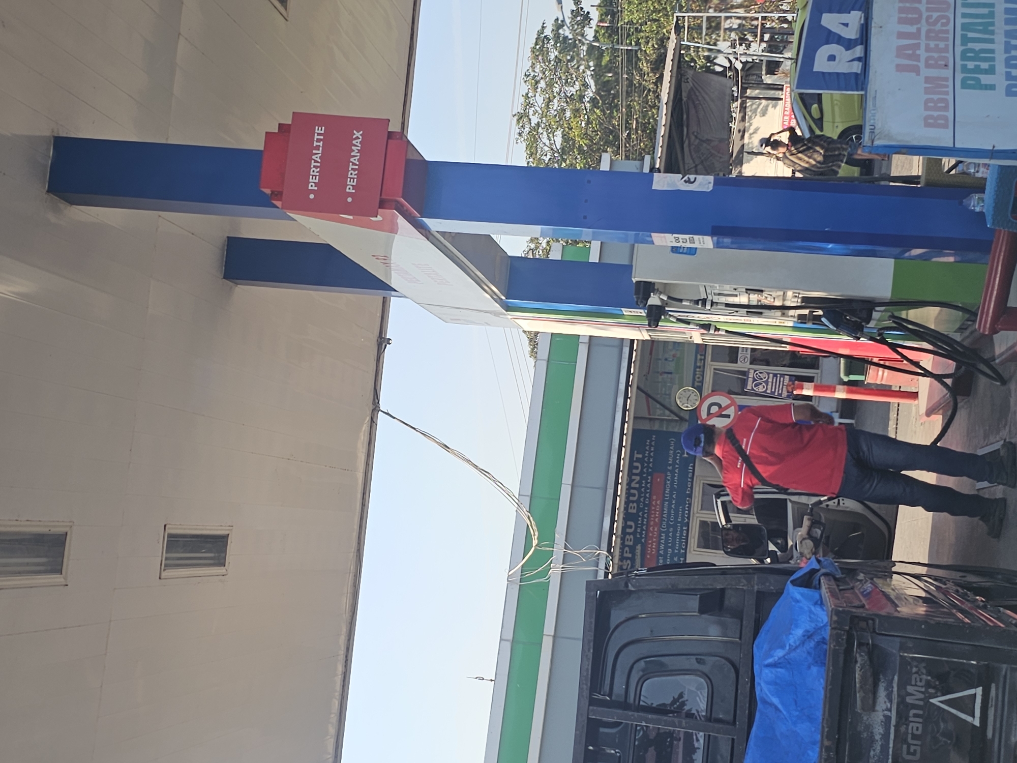 The image shows a gas station in Widang, Indonesia. A man in a red shirt and blue jeans is standing at a gas pump, fueling a black truck. The truck has a blue tarp covering its bed. A gas station attendant in a blue shirt and cap is standing near the truck. A sign above the gas pump reads "PERTALITE" and "PERTAMAX". There are other gas pumps in the background, as well as a small building with green trim. The sky is clear and blue. The overall scene depicts a routine transaction at a gas station.