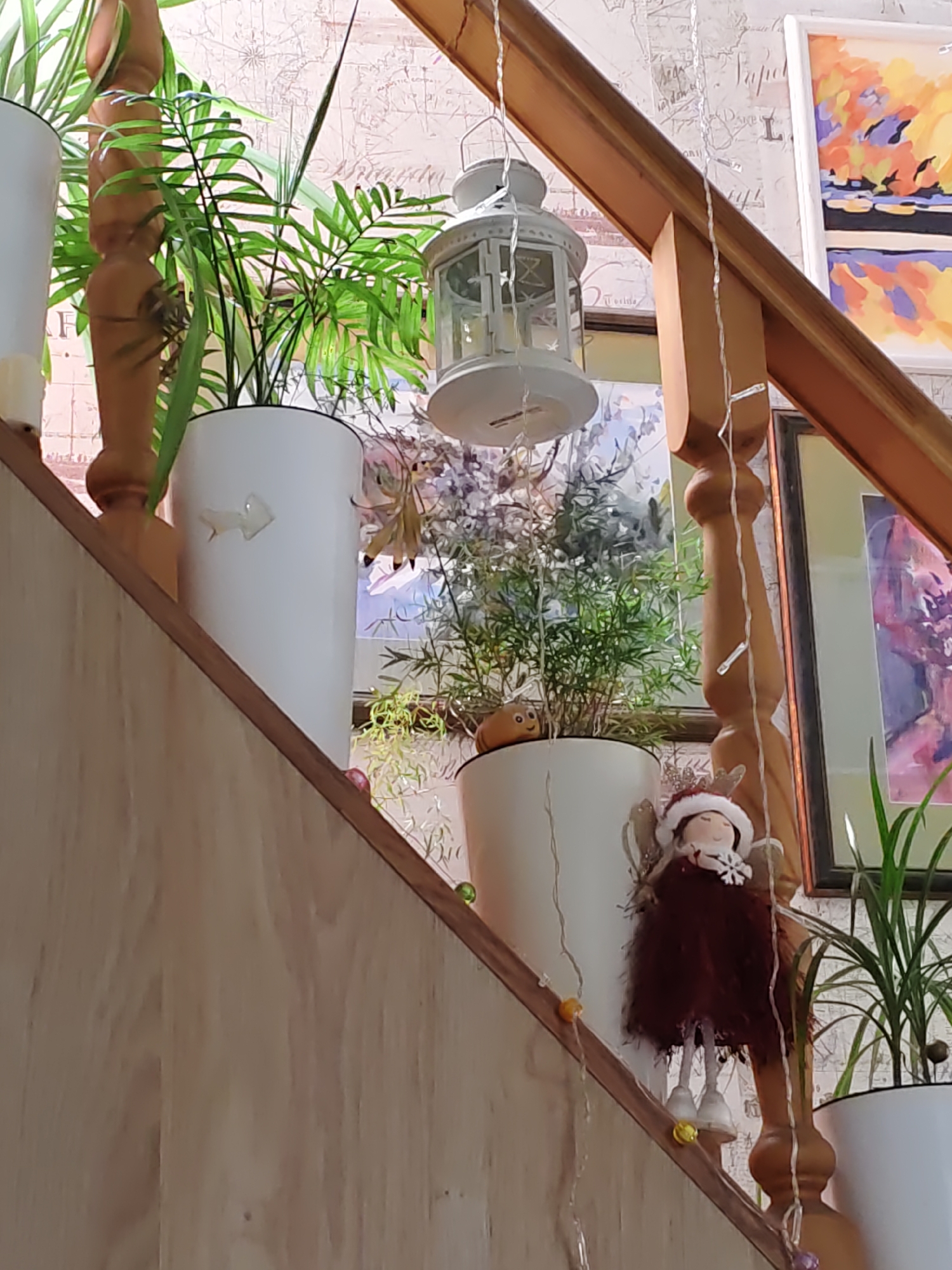 This is a medium-shot, eye-level, indoor photograph taken in Rostov-on-Don, Russia, focusing on a staircase decorated with plants and holiday-themed elements. The staircase features a wooden banister and steps. Potted plants, including a tall palm-like plant in a white pot and a smaller, bushy plant in another white pot, are placed on the steps. A decorative white lantern hangs from the banister. A small angel figurine wearing a red dress and a Santa hat, with iridescent wings, is positioned on a step, adorned with a snowflake on its chest. String lights with small, colorful beads (yellow, green, and purple) are draped along the banister and steps, suggesting a festive atmosphere. In the background, framed artwork with abstract colorful designs is visible on the wall, as is patterned wallpaper. The overall scene conveys a sense of home decoration and seasonal cheer.
