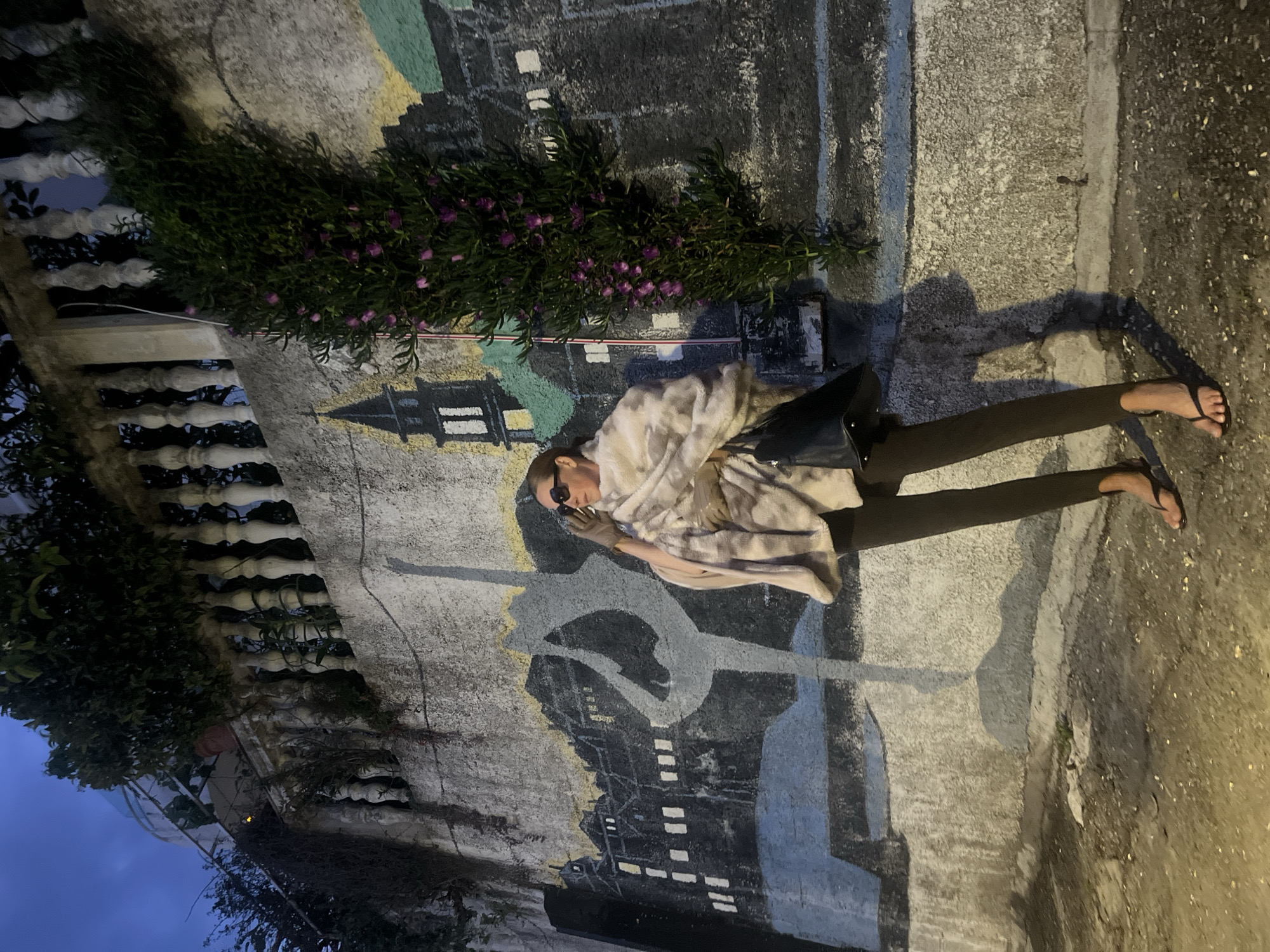 The vertical image captures a person standing on a paved path next to a mural-decorated wall at dusk or night. The person, likely female, is positioned in the lower right portion of the frame. They wear sunglasses, a light-colored, patterned top or shawl, dark leggings, and black flip-flops. They carry a black bag over one shoulder and are seen with their hands raised near their face, possibly interacting with a phone or adjusting their eyewear.
The wall features a painted mural depicting a stylized cityscape with buildings, some with illuminated windows, and a prominent silhouette, possibly a tower, against a dark background. Above the person's head, the mural includes a lighter, abstract design in yellow and light blue. Along the top of the mural, a long planter box holds lush dark green foliage with small purple flowers. To the far left, a white ornate balustrade or railing and more dark greenery are visible against a deep blue evening sky.
Artificial lighting from an unseen source casts strong, elongated shadows of the person and other objects on the ground and wall, emphasizing the late hour. The ground is a rough, concrete-like surface. The scene suggests an urban or semi-urban setting, possibly a pathway in a town like Budva, Montenegro, given the context. No discernible text is visible within the image.
