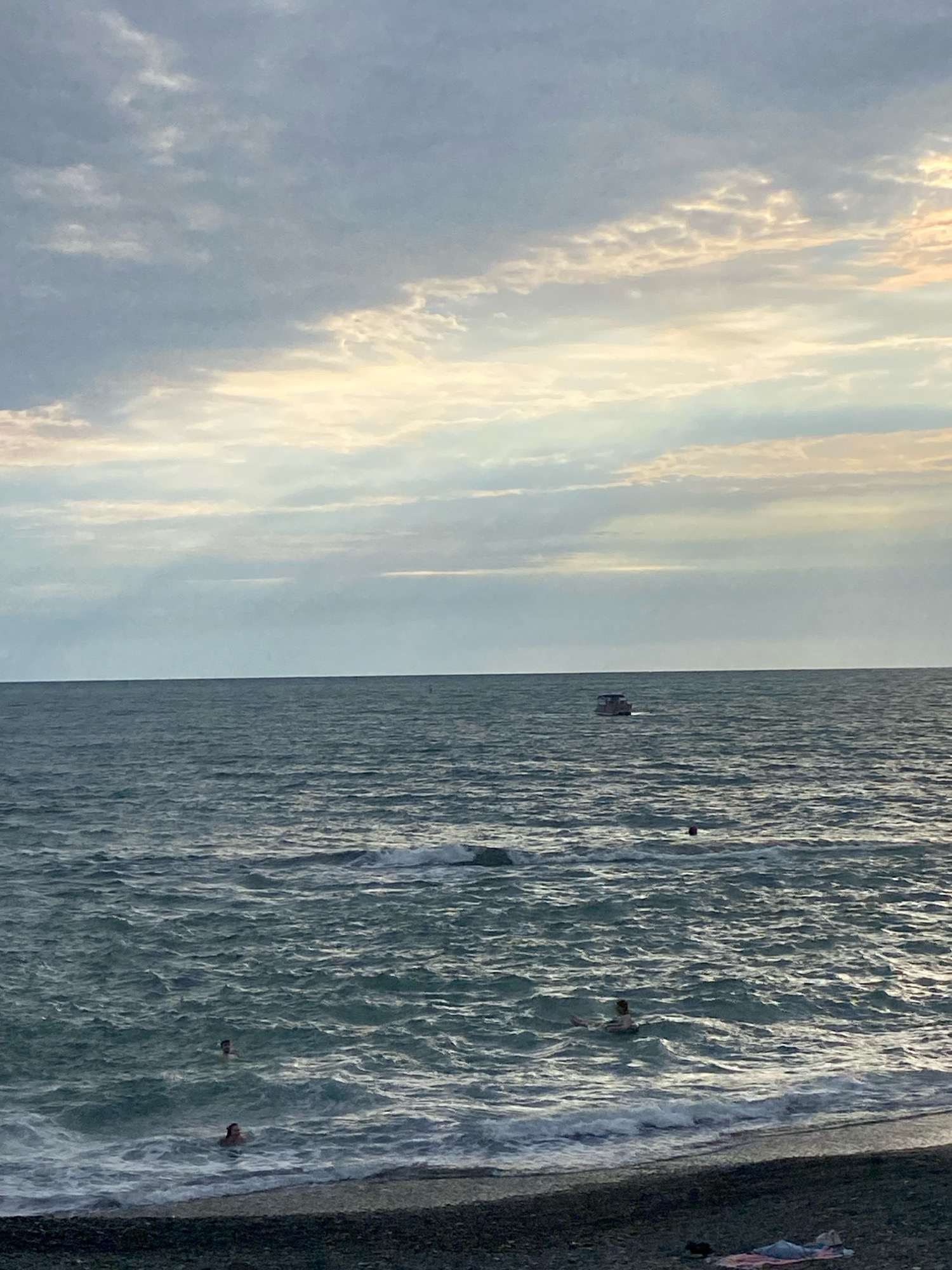 The image depicts a cloudy sky over a choppy sea, with a small boat in the distance. Several people are visible swimming in the water closer to the shore, and one person is lying on a towel on the pebble beach. The scene appears to be a typical beach setting, likely in Sochi, Russia, given the context of the prompt. The sky is filled with a mix of grey and lighter clouds, with streaks of sunlight breaking through in some areas. The sea is a deep blue-green, with whitecaps indicating the rough conditions. The beach is composed of dark pebbles, with a few scattered items on it, including the towel with the person on it. The overall impression is of a somewhat overcast but still active beach day.